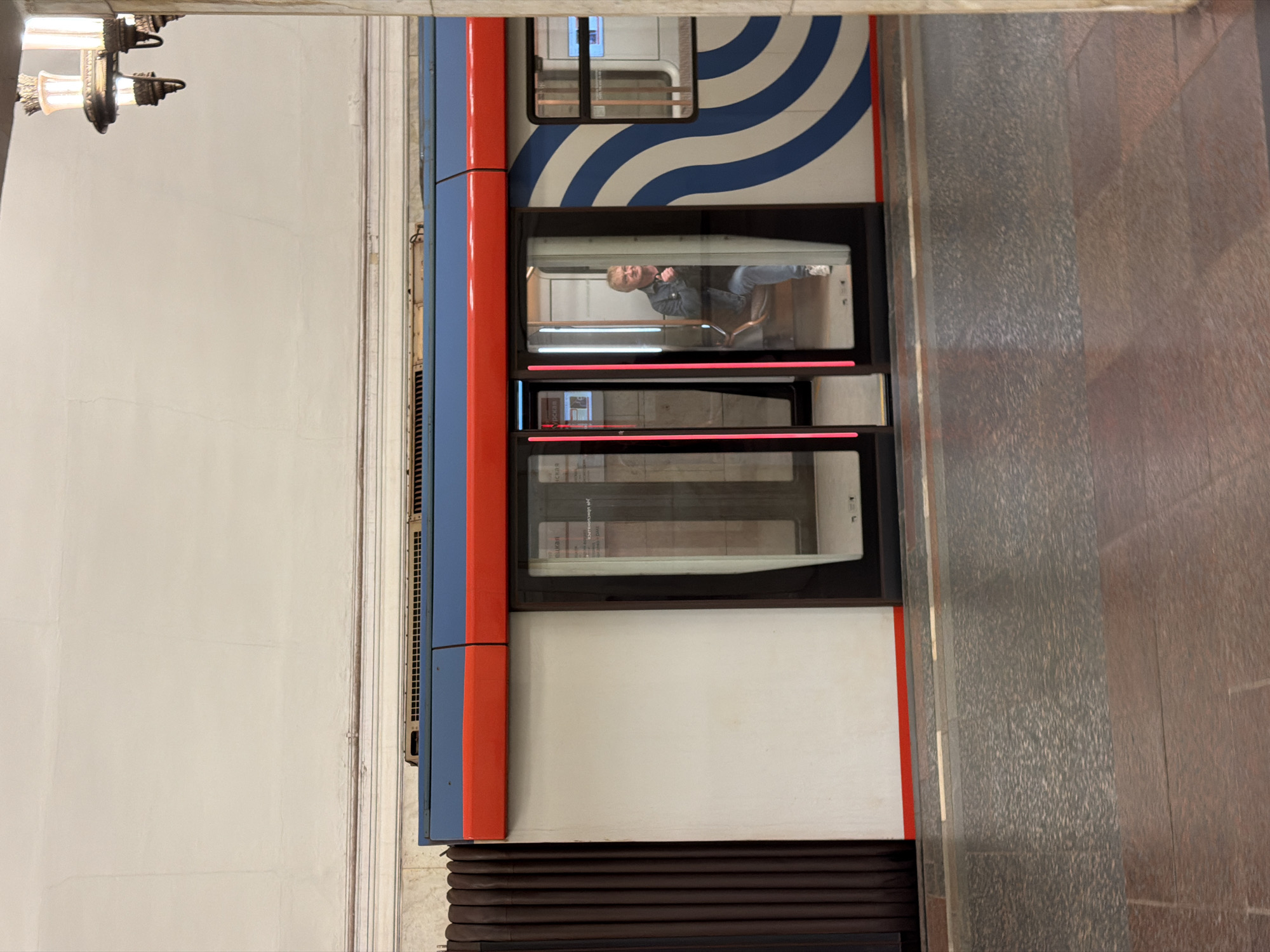 The image captures a section of a Moscow Metro train carriage, distinguished by its blue and white wavy stripes above the windows, with red accents on the carriage's frame. One window reflects a man sitting inside, facing right, wearing a jacket and jeans. The lighting is predominantly indoor artificial illumination. The exterior of the carriage is painted in white and framed by red and blue stripes. To the left, a beige-toned wall is seen, and above, a decorative light fixture hangs. To the right, the Metro station's wall features a gray, textured surface. Below the carriage are dark brown accordion-like connecting sections. The location appears to be a platform inside a Moscow Metro station.