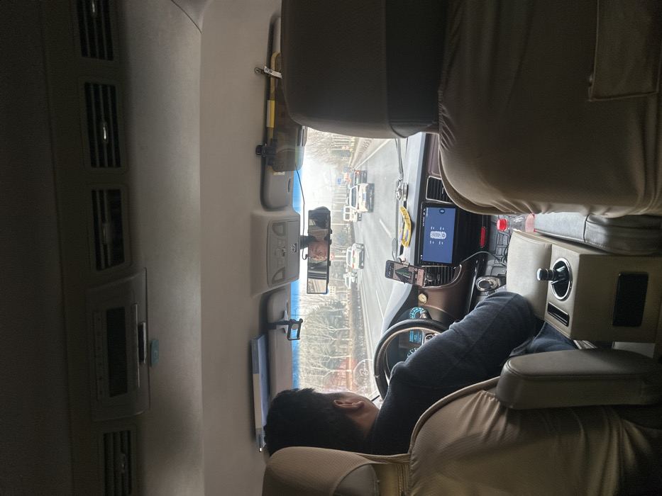 The image depicts the interior perspective from a rear seat of a moving vehicle, likely a van or SUV, traveling on a multi-lane road in Huangdao District, China. A male driver is visible from behind, wearing a dark garment and operating the vehicle with his hands on the steering wheel. His reflection, a male face looking forward, is visible in the wide rearview mirror.
The vehicle's dashboard features a large central display screen showing a white vehicle graphic, potentially indicating tire pressure or parking assist, and a smartphone mounted to the left, displaying a navigation interface. A gear selector knob is located on the center console, which also contains a cup holder holding a red-capped bottle. Overhead, air vents are visible, along with an overhead console unit and a dashcam mounted near the rearview mirror. A long, cylindrical object is secured above the passenger-side sun visor. The driver's seat and the seat in the immediate foreground are covered with light-colored fabric.
Outside the vehicle, a multi-lane highway is visible with moderate traffic, including several white and light-colored cars and a white van, all moving in the same direction. The roadside features a line of deciduous trees, currently bare, suggesting a colder season. The sky appears overcast or hazy, limiting visibility of the distant horizon.