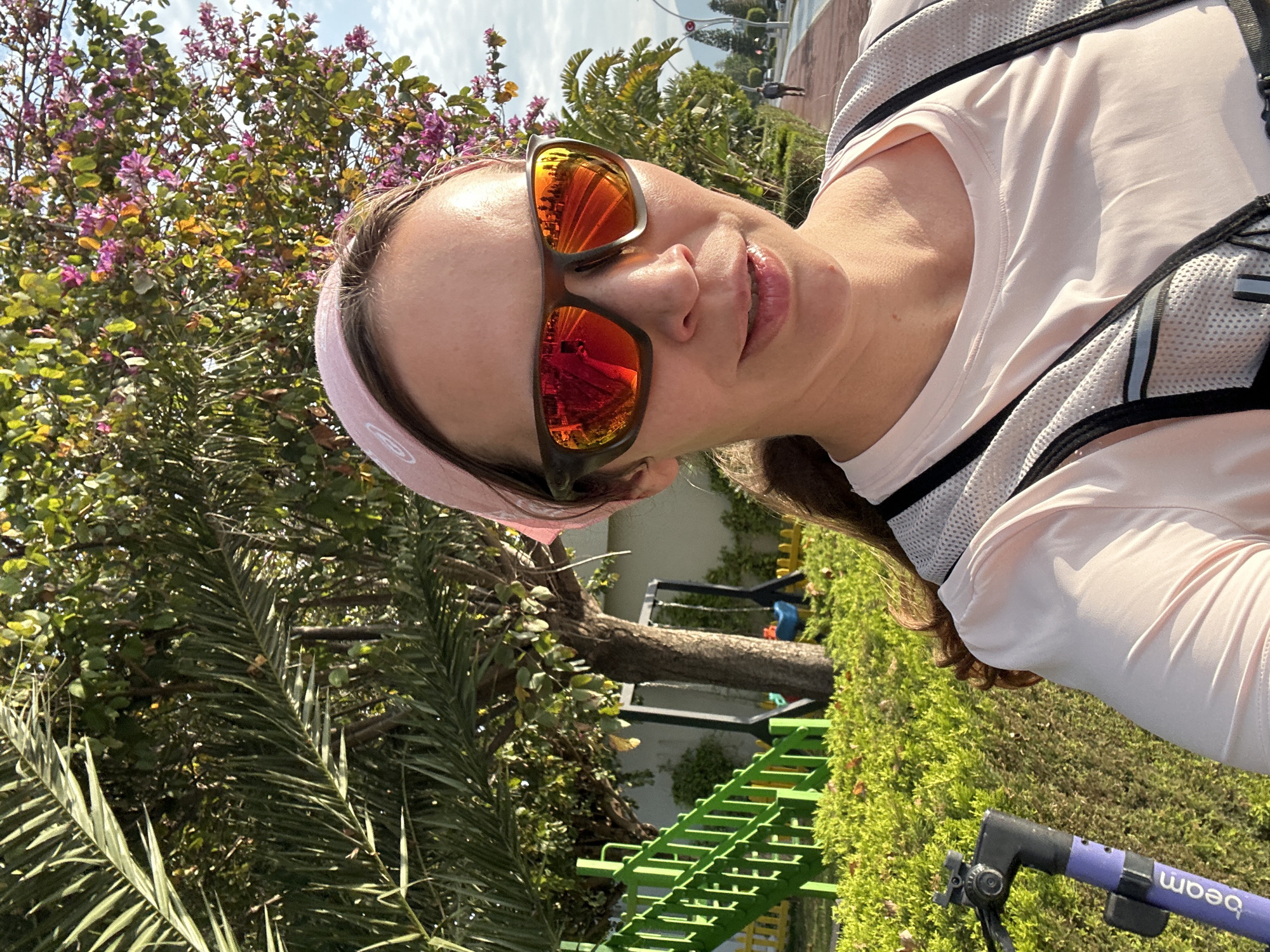 A woman wearing a pink headband and reflective sunglasses is posing for a selfie outdoors. She is wearing a light pink athletic shirt and a mesh backpack. A palm tree and a tree with pink blossoms are visible to her left. A playground with green metal stairs is visible in the background, along with a green hedge. The picture appears to be taken at a park or public space in Fethiye, Turkey, as indicated by the "beam" logo on a purple object near the bottom of the frame, which likely belongs to a Beam e-scooter available in Fethiye.