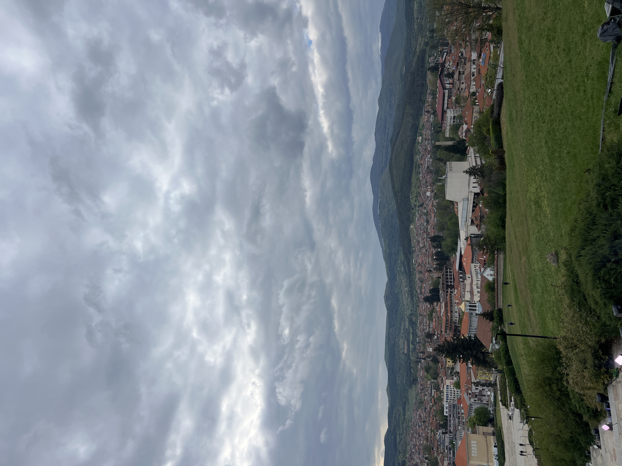 The media file presents a panoramic view of Panagyurishte, Bulgaria, from an elevated vantage point, likely a hillside or park. The image is oriented vertically but depicts a horizontal landscape.
The sky, filling a significant portion of the upper frame (left in the original image), is heavily overcast with a mass of grey and white clouds, indicating a cloudy or partly cloudy day with diffuse light. No direct sunlight is visible.
In the mid-ground, a sprawling town stretches across the frame, nestled at the base of a range of green, rolling mountains or hills that form the distant horizon. The town is characterized by numerous buildings, many featuring traditional reddish-brown tiled roofs. Some larger, more modern buildings with lighter-colored facades and flat roofs are also visible among the residential areas. The town appears to follow the contours of the valley or a river.
The foreground (right side of the original image) consists of a well-maintained grassy slope, with some trees and bushes. A paved walkway or path is discernible in the lower right, lined with what appear to be streetlights or garden lights. A few small, indistinct figures of people can be seen walking on the grass or path, but their details are not clear.
The overall impression is of a calm, cloudy day over a scenic town surrounded by nature. There is no visible text in the image.