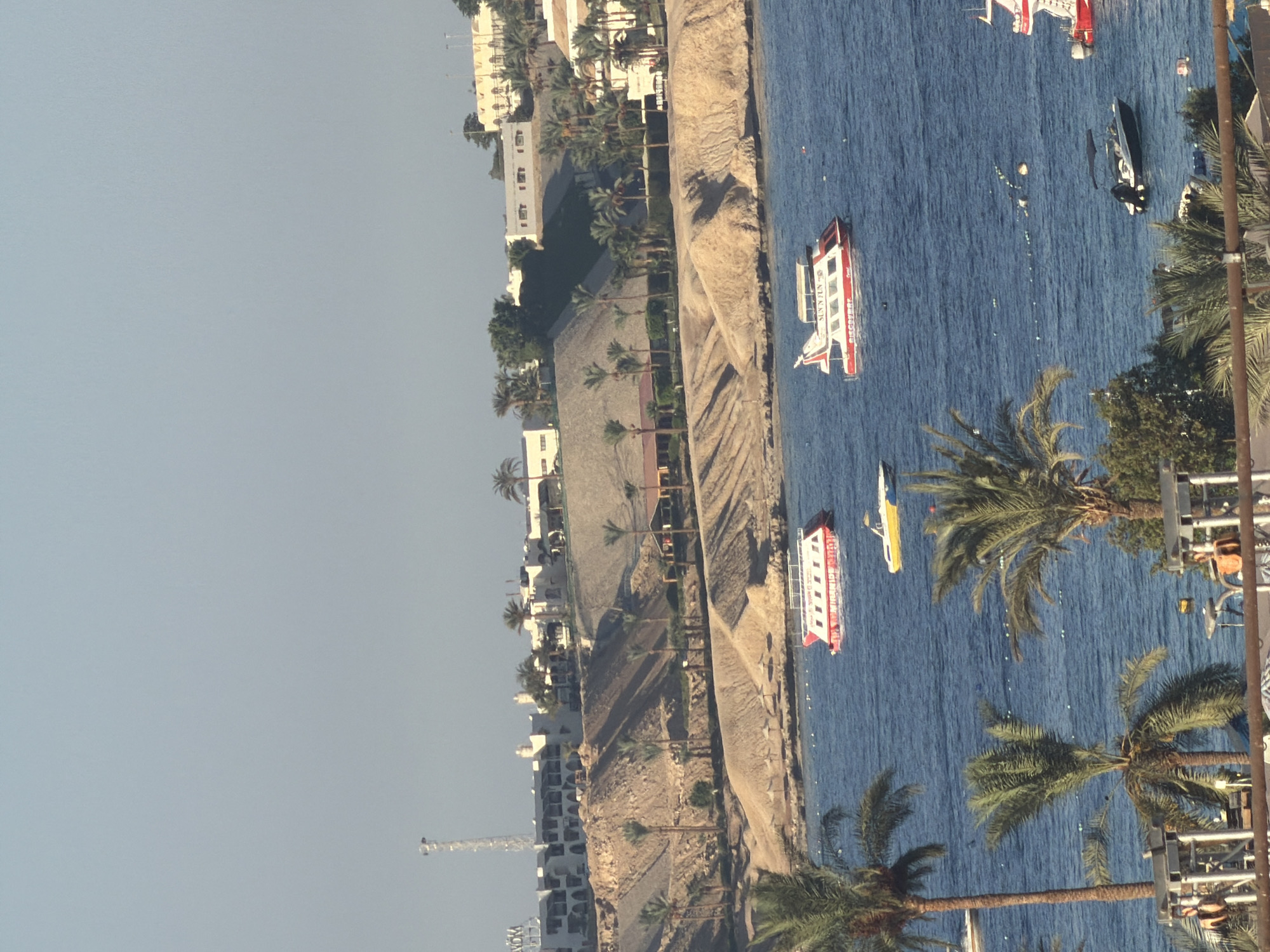The image captures a coastal scene in Sharm El Sheikh, Egypt, with a view of the Red Sea and a hillside dotted with resort buildings and palm trees. In the foreground, a portion of a resort structure with metal railings and seating areas is visible. The sea is a vibrant blue, with several boats on its surface. Two prominent red and white tour boats, one labeled "SUN'N FUN," are anchored or moving slowly. A smaller yellow boat is also present, along with a dark-colored motorboat. In the background, a beige-colored hill rises, featuring terraced resort buildings with white facades and dark rooftops. Palm trees are scattered across the hillside and along the coastline. A communication tower can be seen on the far left of the hillside. The sky is a pale blue, indicating a clear day. The overall impression is of a sunny resort destination.