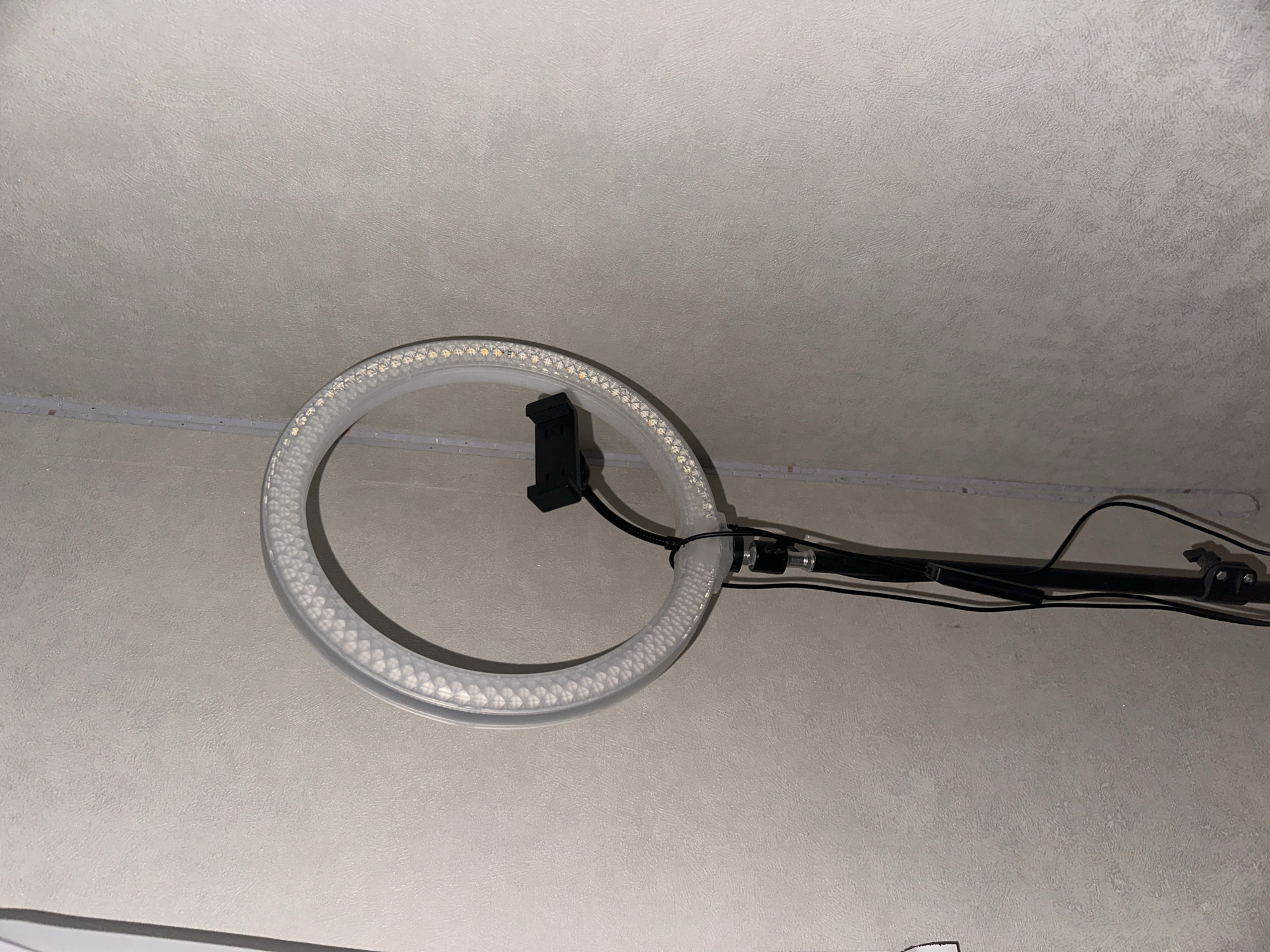 A ring light is positioned in front of a textured, light-colored wall. The ring light is circular with many small LED lights around its circumference. A black phone mount is attached to the center of the ring light. Cables are visible running down from the ring light to a black stand. The overall environment appears to be indoors.