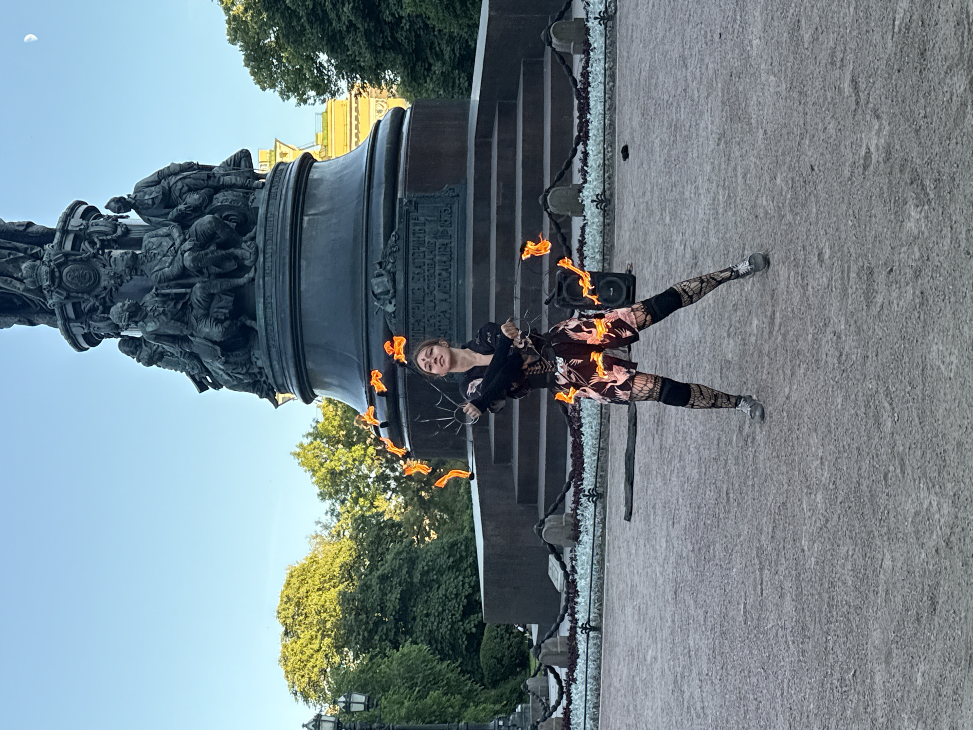 A young woman with face paint performs fire spinning in front of a large, ornate monument in Palace Square, Saint Petersburg, Russia. She is dressed in a black top, patterned shorts with pink cranes and black fishnet stockings. Several fiery props are visible around her, some held in her hands and others in motion. The monument, which appears to be the Alexander Column, is a prominent feature in the background, with its bronze sculptures and intricate details illuminated by the setting sun. Trees and a lamppost are also visible, contributing to the outdoor urban setting. The sky is clear with a hint of a crescent moon.