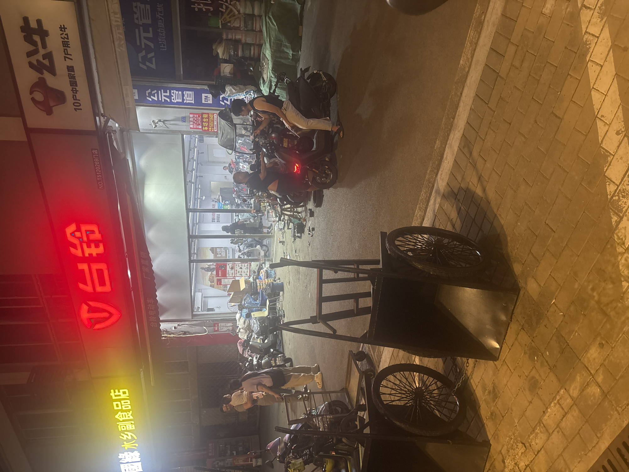 The image shows a street scene at night in the Suzhou Industrial Park, China. In the foreground, two large, black, wheeled carts are visible. One cart has a large black body and two large spoked wheels, while the other is partially obscured but appears similar. The carts are positioned on a brick-paved sidewalk.
In the midground, a store with a bright red neon sign that reads "台铃" (Tailing) is prominent. To the left of the "台铃" sign, a yellow neon sign reads "國緣 水鄉副食品店" (Guoyuan Water Town Food Store). The store appears to be an electric scooter shop, with several electric scooters parked both inside and outside. A man and a woman are standing near the entrance of the store. The man is holding a young child.
Further back, more electric scooters are visible, along with stacks of goods and bags, suggesting a commercial area. People are milling about and interacting with the scooters. A blue sign on the store reads "公元管道" (Gongyuan Pipe). Another sign above the store entrance features a logo and text that appears to be advertising "公牛" (Gongniu) products, possibly related to household appliances or electronics. The overall environment suggests a busy commercial street at night.