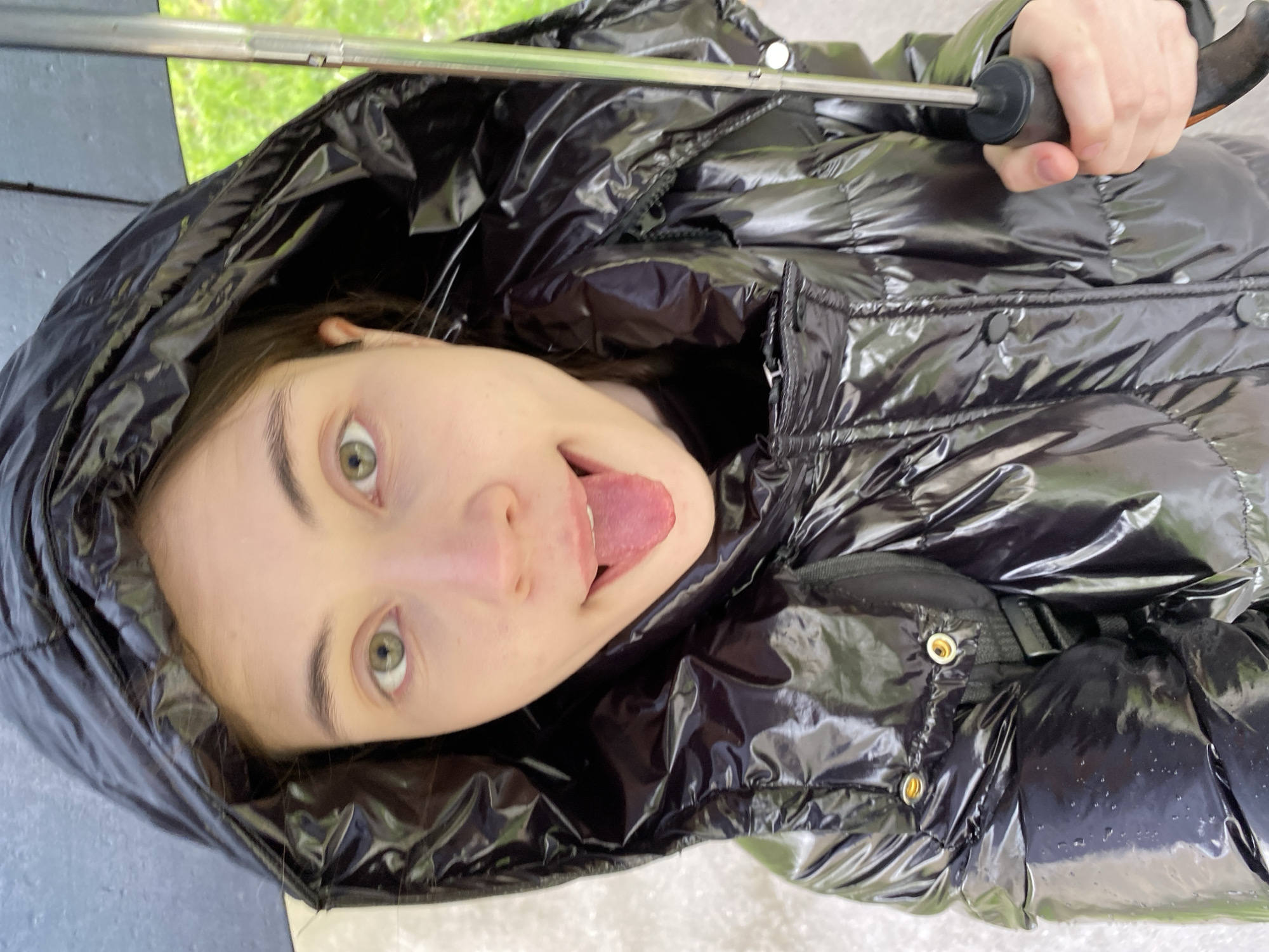 A young woman is pictured at a close angle wearing a black, puffy coat with a hood up. She is sticking her tongue out and looking at the camera. She is holding a black umbrella with a silver shaft. The environment appears to be outdoors. It is not possible to determine the specific location in Votkinsk, Russia from the provided image.