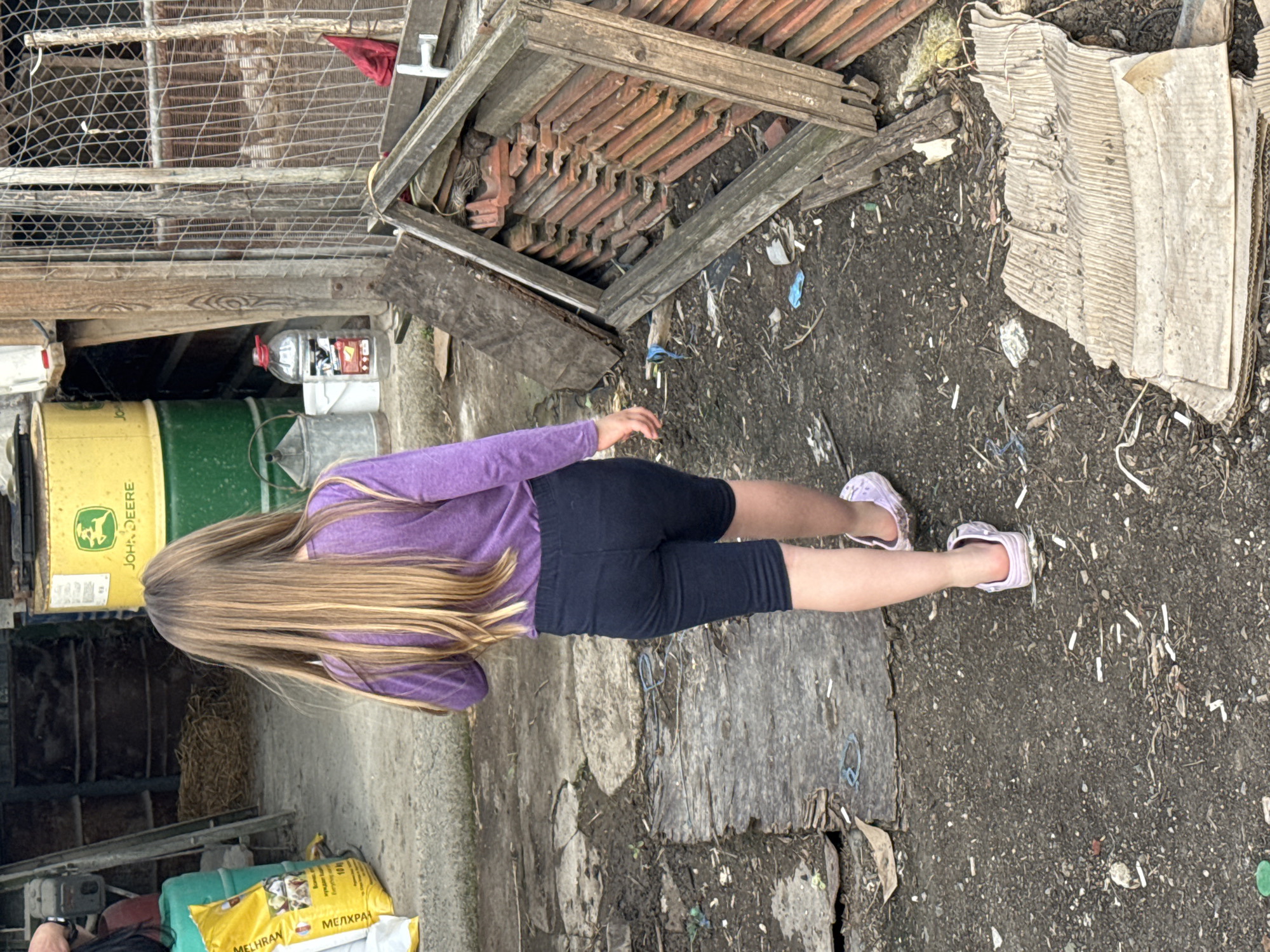 A person stands outdoors in Sarnevo, Bulgaria. The person, wearing a purple shirt, dark shorts, and light purple sandals, faces away from the viewer. The environment contains discarded tiles, wood, cardboard, and other refuse. A John Deere barrel and a bag labeled "MEHRAN" are visible. A metal cage is visible in the background.