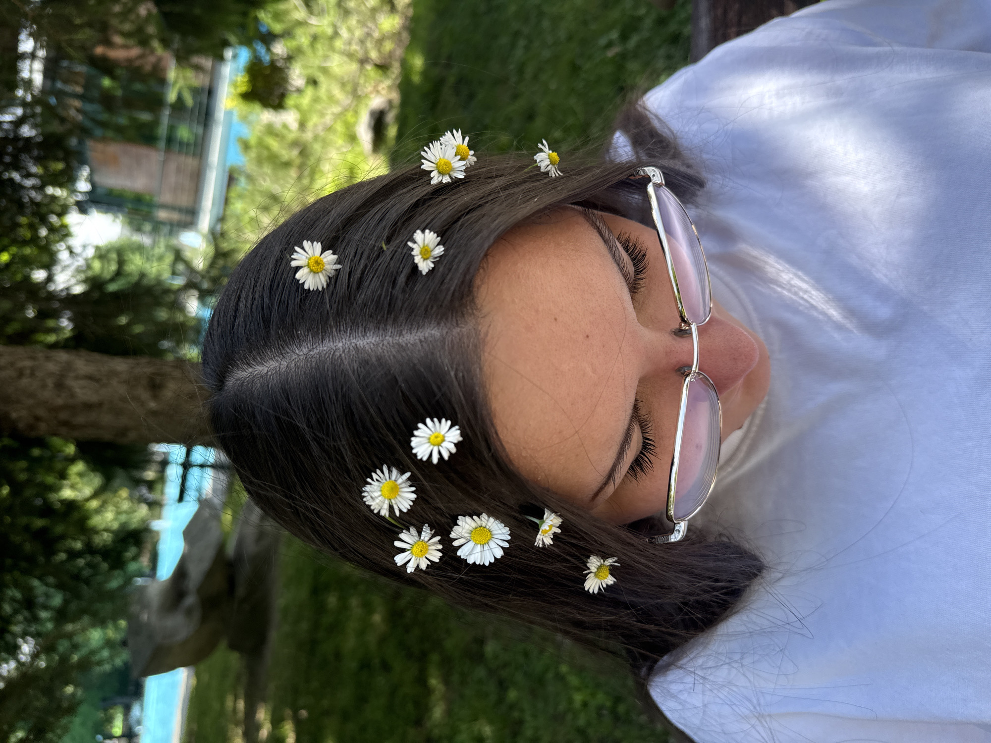 A young woman with dark hair is shown from a high angle, looking down. Several small white daisies with yellow centers are placed in her hair. She is wearing a white t-shirt and reflective eyeglasses. The background shows a blurry outdoor setting with trees, green grass, and a body of water, possibly a pool, with a grey sculpted element on its edge. The scene appears to be in a park or garden area. The exact location in the city of Plovdiv, Bulgaria cannot be determined from the image alone.