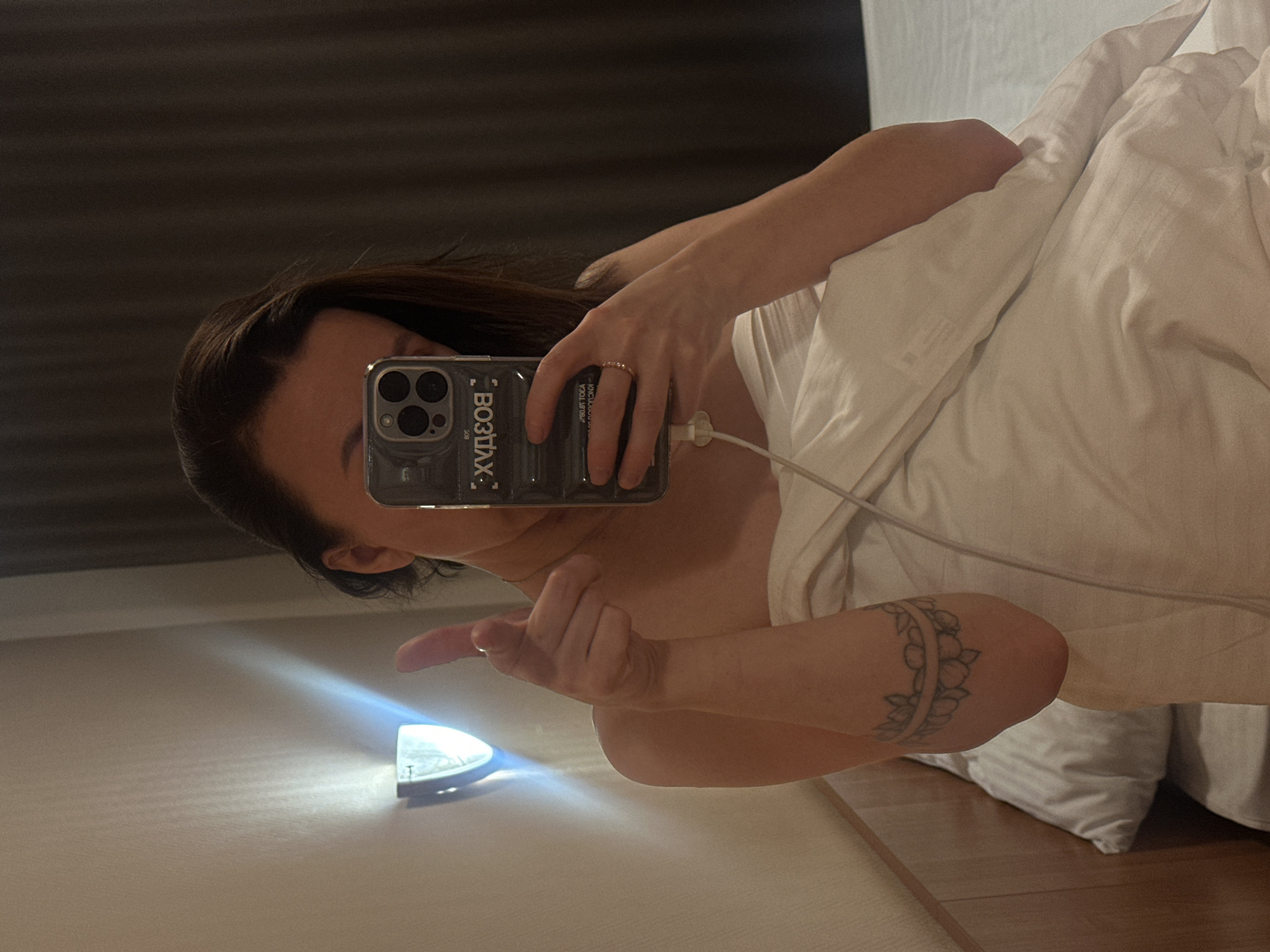 The image captures a selfie taken in a mirror or reflective surface, featuring a person (likely female) partially covered by white bedding, presumably in a bedroom or hotel room in Perm, Russia. The person is holding a smartphone in their right hand, using it to capture the reflection. A thin ring is visible on their right ring finger, and a white charging cable is plugged into the phone. The phone itself, an iPhone model with a triple camera setup, is in a clear case that displays reversed Russian text: "ВОЗДУХ" (VOZDUKH, meaning "AIR"), "ЫЛХЕЧ ЕВОВПОТ" (reversed for "ТОПОВЫЕ ЧЕХЛЫ" or "TOP CASES"), "80.1" (reversed for "1.08"), and "ЙО!" (reversed for "OY!," an interjection).
The person's dark hair frames their face, which is partially visible. Their left arm is extended, with the index finger pointing towards a small, bright, illuminated object (possibly a portable lamp or flashlight) positioned on a light-colored surface below. This light casts a strong beam upwards. A band-style tattoo with floral or leafy motifs is visible on the person's left forearm/bicep area. The background behind the person's head appears to be a dark, plain wall or headboard. The overall lighting is dim, suggesting it might be nighttime or a poorly lit interior. The scene conveys a casual, private moment.