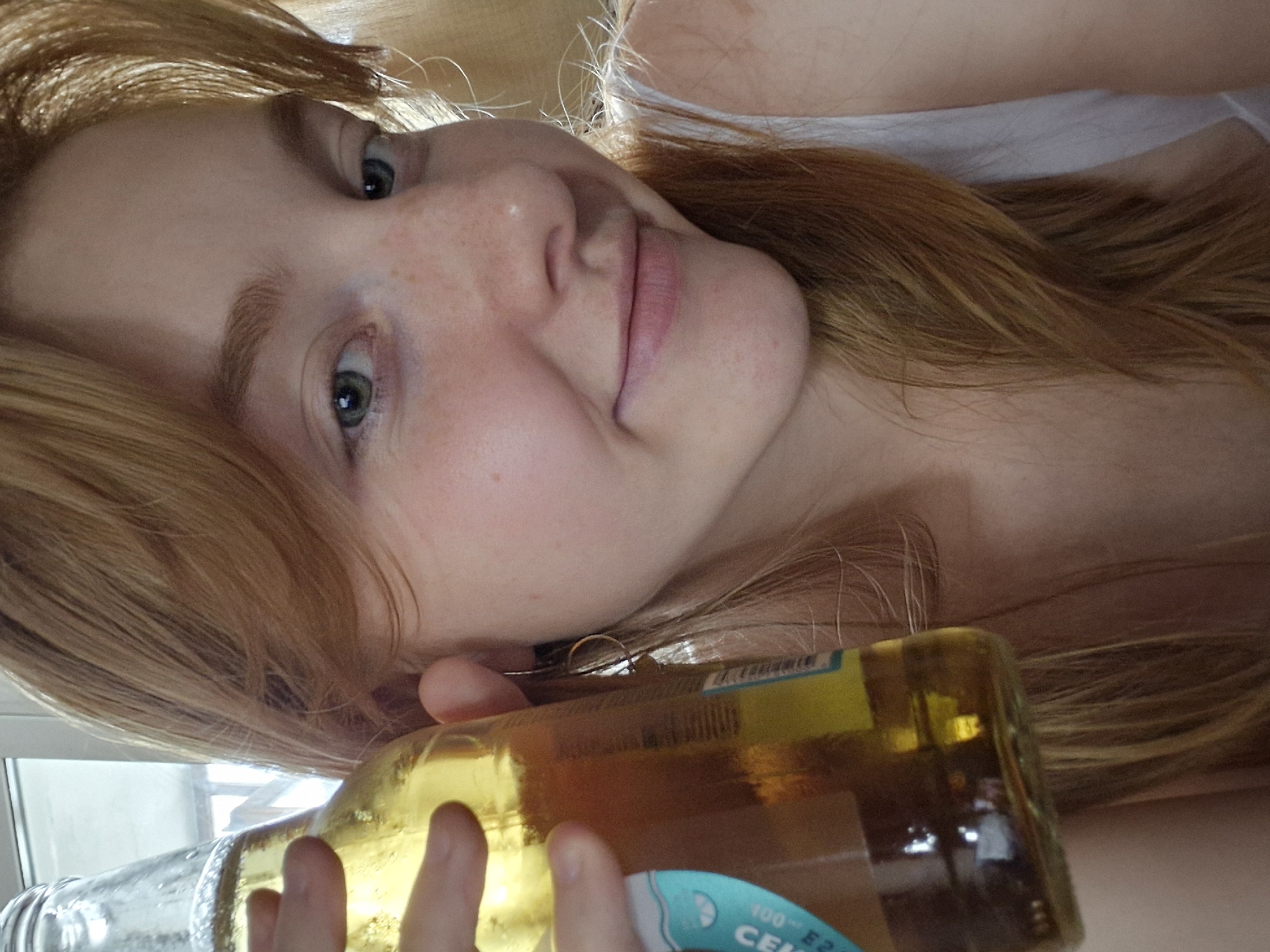 A young woman with auburn hair and freckles is holding a bottle of beer up to her face, with her left hand supporting the bottom of the bottle. She is looking towards the camera with a slight smile. The background is blurred but appears to be an indoor setting with a window and natural light. The image is a close-up shot, focusing on the woman's face and the bottle. There is no specific indication of the location being in Yekaterinburg, Russia.