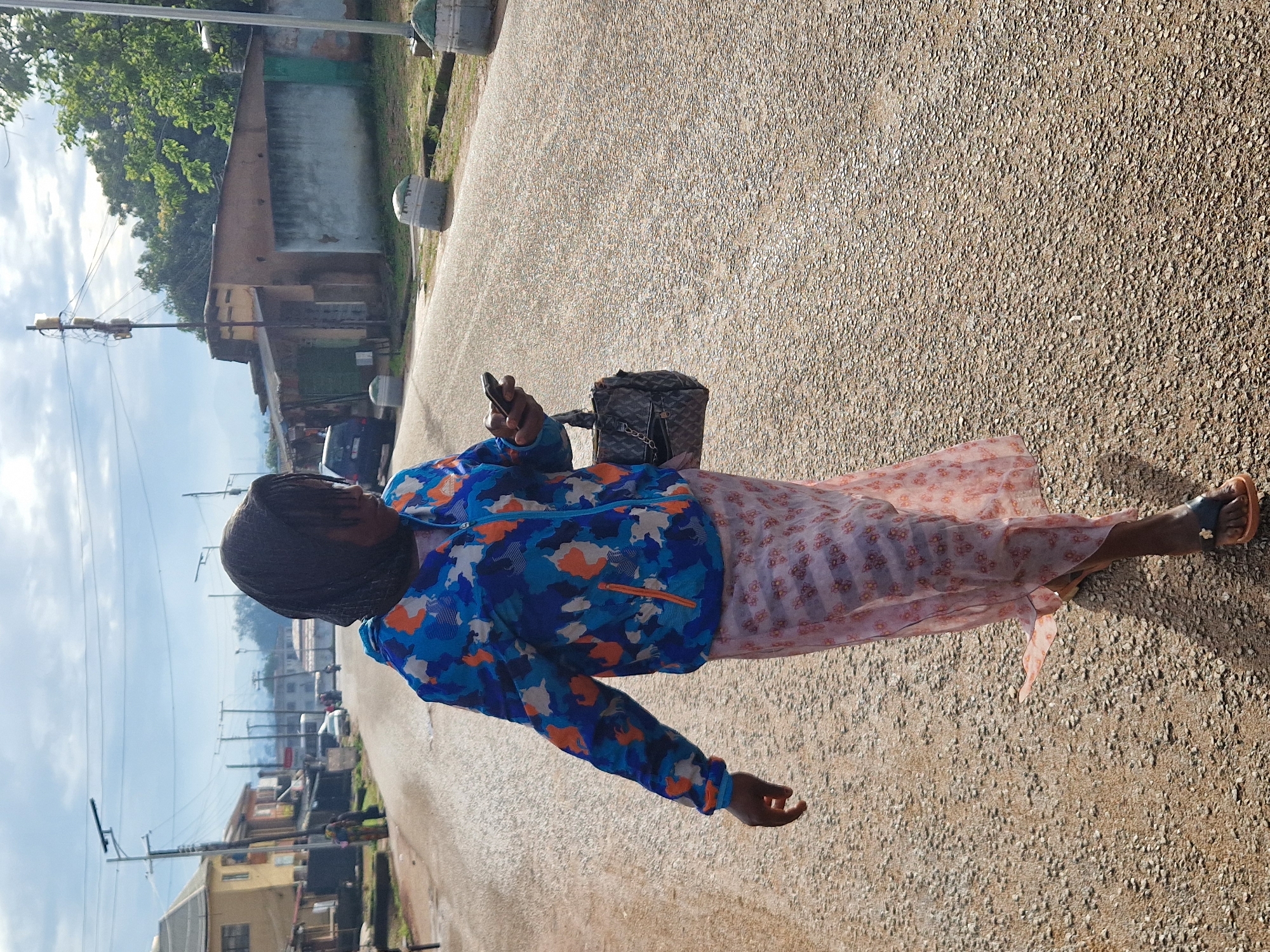 A person walks down a dirt road in Kabong, Nigeria. The person is wearing a blue, orange, and white camouflage jacket over a long pink floral skirt and black and orange sandals. They are holding a black smartphone in their right hand and carrying a dark-colored handbag over their left shoulder. The road is lined with buildings, some of which appear to be residences or small businesses, and utility poles with electrical wires. The sky is overcast with some patches of blue.