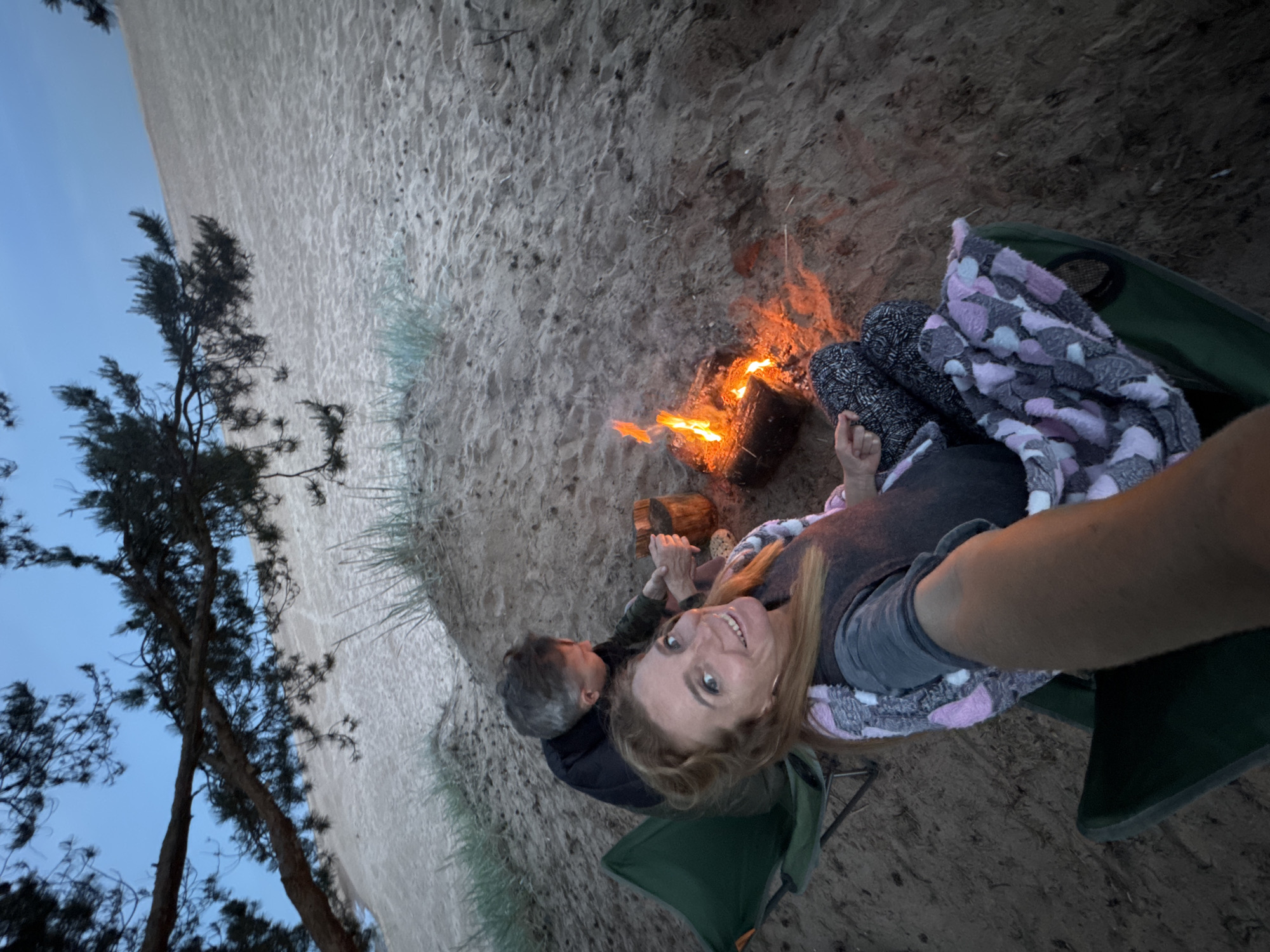 An overhead-angle selfie captures two individuals seated around a small campfire on a sandy dune in Bolshaya Izhora, Russia, during twilight. In the foreground, a woman with light brown hair, wearing a dark top, patterned pants, and a pink, grey, and white patterned blanket, smiles directly at the camera while holding the camera with her right arm, which is partially visible. She sits on a green folding camping chair. To her left and slightly behind, a man with short grey hair, wearing a dark jacket, sits on another green camping chair, facing away from the camera. He appears to be tending to a small pot or container placed next to a burning campfire. The campfire, centrally located, consists of a small log with visible orange flames. The immediate ground is sandy, scattered with sparse, light green vegetation. In the background, a large, expansive sand dune slopes upwards under a pale blue sky, with dark, pine-like tree branches extending from the upper left and right across the frame.