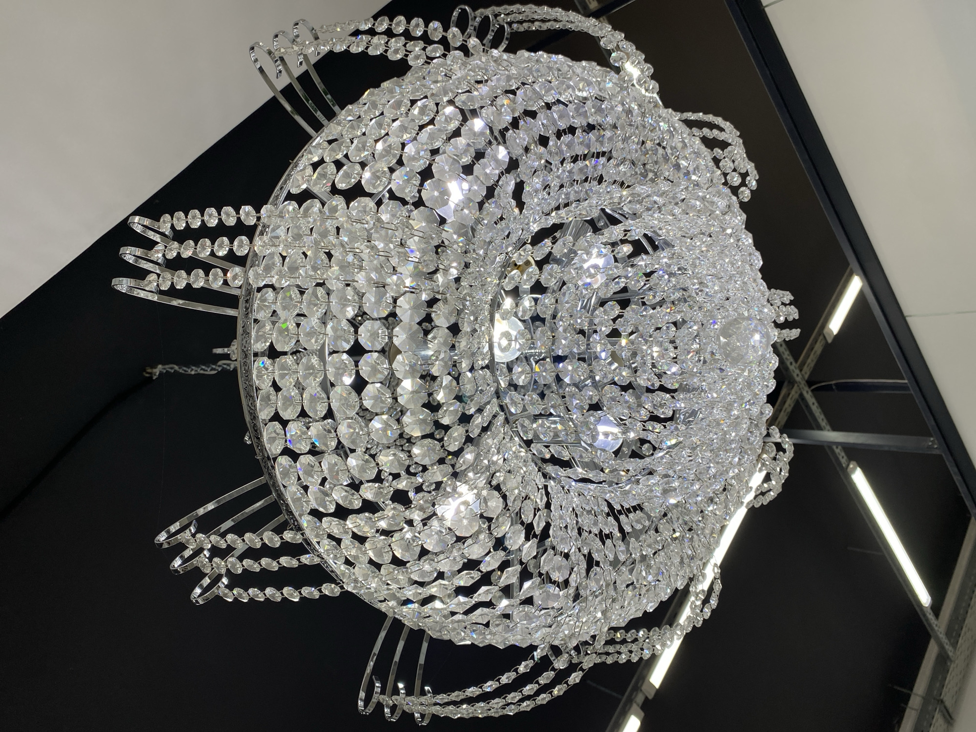 This media file features a large, elaborate crystal chandelier, captured from a low-angle perspective looking upwards. The chandelier is the central subject, dominating the frame with its intricate design and sparkling appearance. It is composed of countless clear, faceted crystal beads strung together in multiple cascading layers, forming a voluminous, spherical or globe-like shape. The crystals brilliantly reflect and refract light, creating numerous bright highlights and subtle rainbow prisms across their surfaces. The framework supporting the crystals appears to be made of polished silver-toned metal, with curved elements and thin wires visible through the crystal strands.
The background primarily consists of a dark ceiling, providing a strong contrast to the luminous chandelier. Parts of a lighter-colored ceiling or wall are visible in the upper sections of the image, separated by dark linear elements that suggest structural beams or framing. On the right side, in the deeper background, there are indications of an indoor commercial or industrial setting, with visible fluorescent light fixtures and metal racking or support structures.
No people are visible, and no specific activity is taking place; the image is a static shot of the chandelier. The lighting suggests an indoor setting with artificial illumination. There is no visible text in the image. The location context provided is Votkinsk, Russia, implying this is an interior shot within that city.
