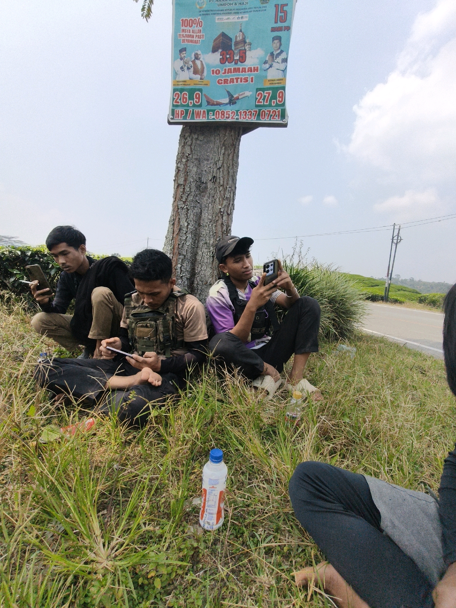 In the city of Pasar Minggu, Indonesia, a group of four young men are seated on a grassy slope next to a tree with a large advertisement attached to it. The advertisement promotes Umrah and Hajj travel packages. Two men are squatting, while the other two are sitting cross-legged. All four men are looking at their mobile phones. One man is wearing a tactical vest over a brown t-shirt, and another is wearing a purple t-shirt with a black vest. A bottle of water is placed on the ground in front of them. The background shows a road and a lush, green landscape, likely tea plantations, under a partly cloudy sky.