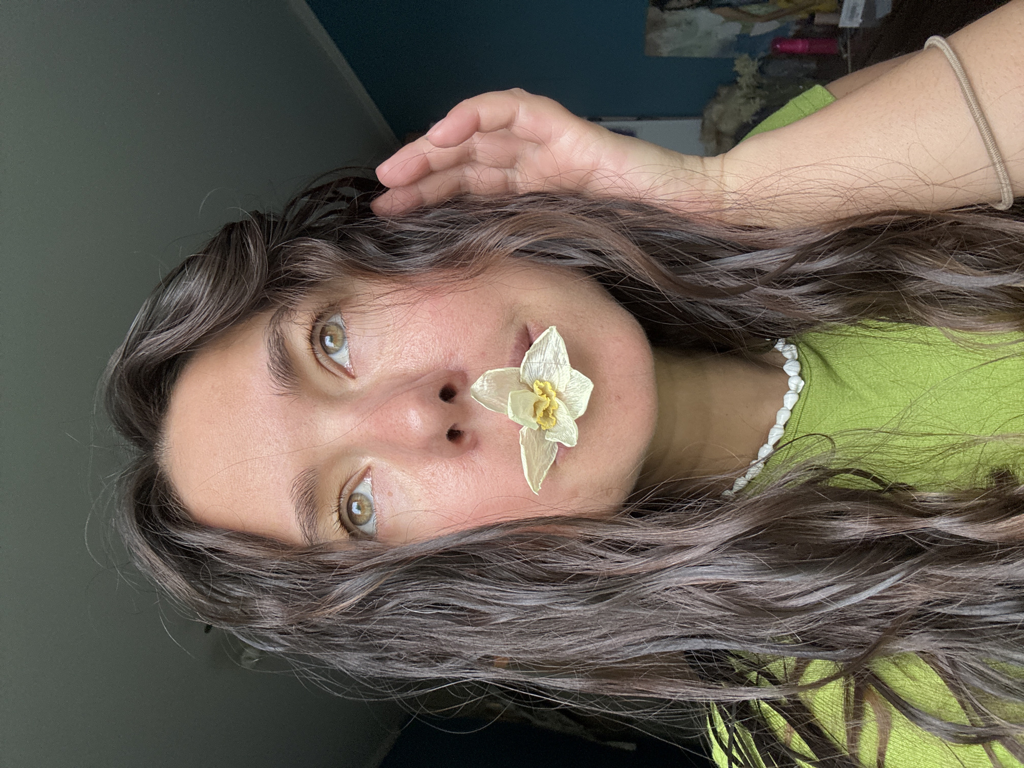 A young woman with long, dark brown hair is positioned in front of a green-blue wall. She is wearing a light green shirt and a white beaded necklace. A dried white flower is placed between her nose and upper lip. Her left hand is raised to her hair, with her fingers touching her hairline. The location is not discernible from the image. The city of Kazan, Russia is not discernible from the image.