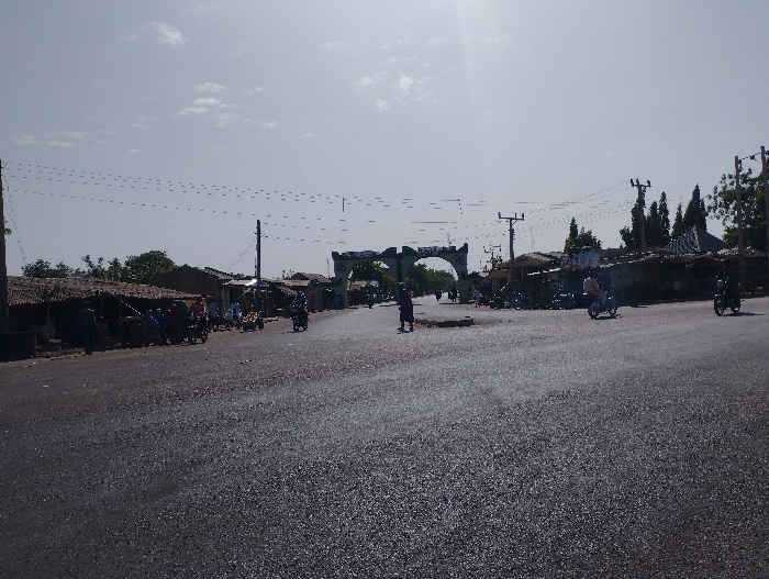 The image shows a paved road with a decorative archway in the distance. There are several people walking along the road, as well as some motorcycles. The road is lined with buildings and trees. There are power lines overhead, and the sky is bright and clear. This is a view of a road in Kumo, Nigeria, located at latitude 10.05 and longitude 11.2. The road appears to be well-traveled and the buildings on either side are made of brick and concrete. The image was taken from a low angle, with the camera facing the archway. The focus is on the road and its surroundings, and there is little detail visible in the people or objects in the distance.  The image likely shows a street scene during the day time.