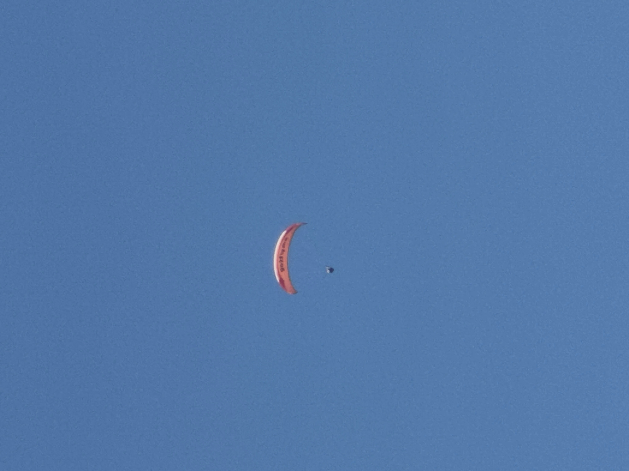 A paraglider is captured in mid-air against a clear blue sky. The paraglider, a vibrant orange with red lettering, is open and suspended above a small, dark figure, presumably the pilot. The paraglider is positioned slightly to the left of the frame's center and appears to be in gentle descent. The environment is a vast expanse of uninterrupted blue sky, with no visible landforms or cityscapes. The image appears to be taken from below, looking upwards. The paraglider has the words "Go2Fly" visible on its surface. The overall impression is one of a serene and solitary flight. The image does not provide any specific information regarding the location in Boreti, Montenegro.