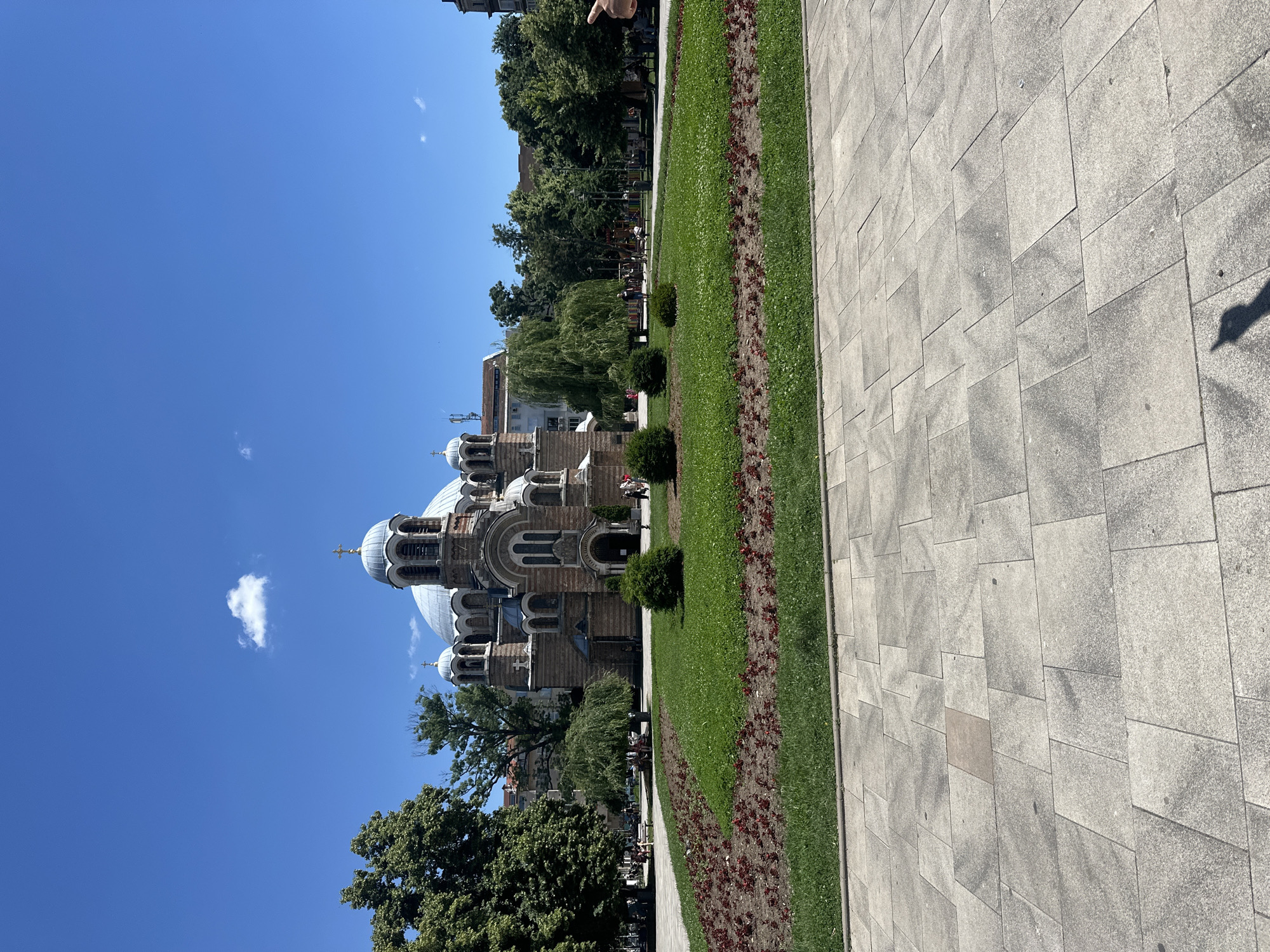 The image depicts an exterior view of a grand cathedral with multiple domes and intricate architectural details, possibly in an Orthodox style. The cathedral is situated in a city square, with a well-maintained lawn and flowerbeds in the foreground. Several trees, including a large willow, are scattered around the square, and a paved walkway is visible in the lower portion of the image. A few people are scattered throughout the scene, some walking and others sitting on benches. The sky is a clear, bright blue with a few scattered clouds, indicating a sunny day. The overall atmosphere is serene and suggests a public space in an urban setting.
