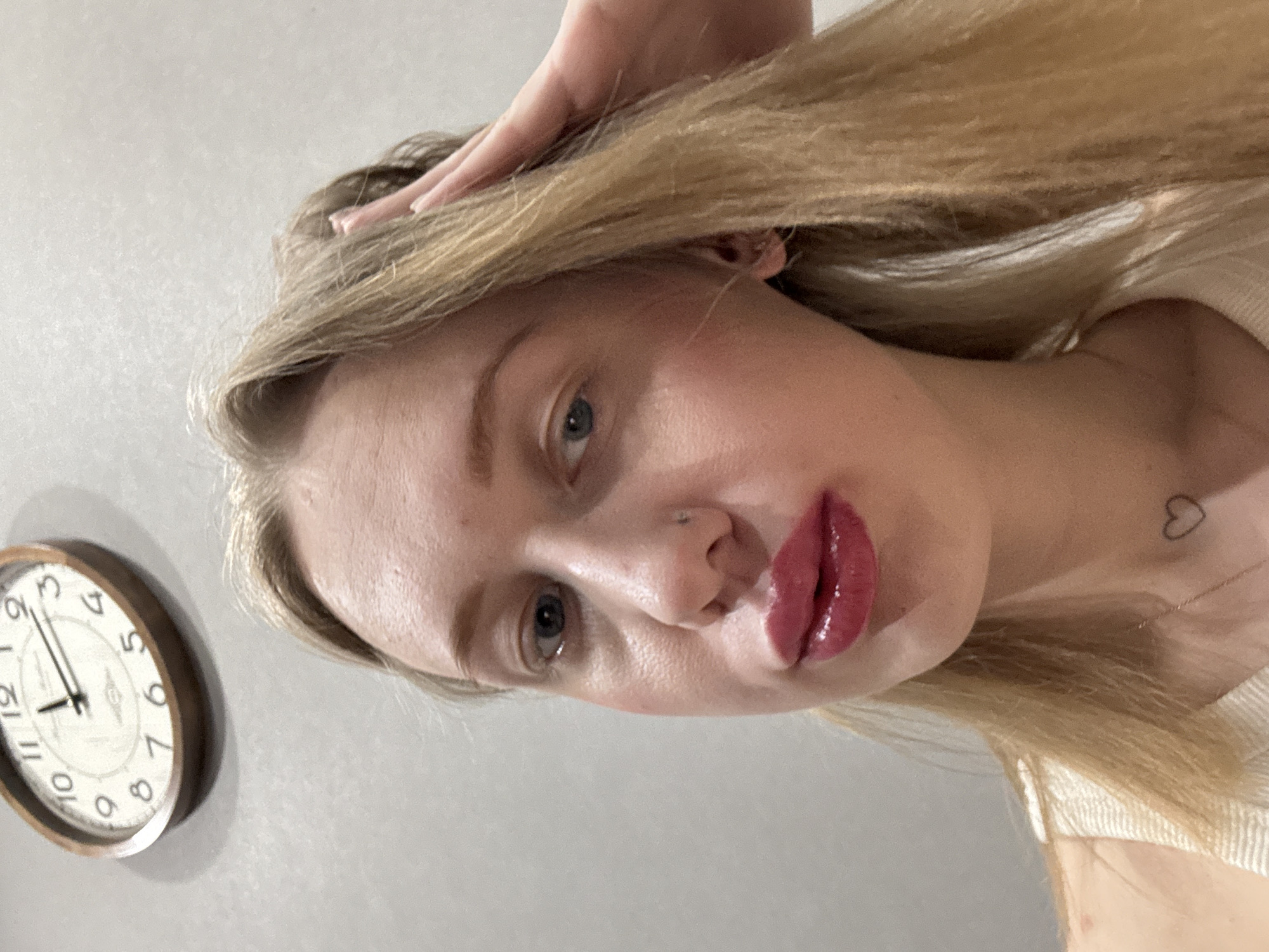 This image depicts a close-up, upward-angled self-portrait of a young adult female, identified as being in Rostov-on-Don, Russia. The subject is a fair-skinned individual with long, straight blonde hair and blue eyes, looking directly into the camera. Her lips are coated with a glossy, dark pink or red lipstick. A small silver-colored stud is visible in her left nostril. Her left hand is raised, with fingers touching the blonde hair on the right side of her head. She is wearing a light-colored, possibly white or cream, ribbed garment, likely a tank top or camisole. A thin gold-colored necklace is visible, and a small, black outline of a heart is tattooed or drawn on the lower right side of her neck/upper chest. The background consists of a plain, light grey wall. Mounted on the upper left portion of the wall is an oval-shaped clock with a light wooden frame and a white face with black Arabic numerals. The clock hands indicate a time approaching the hour, with the minute hand at the eleven and the hour hand between the one and two.