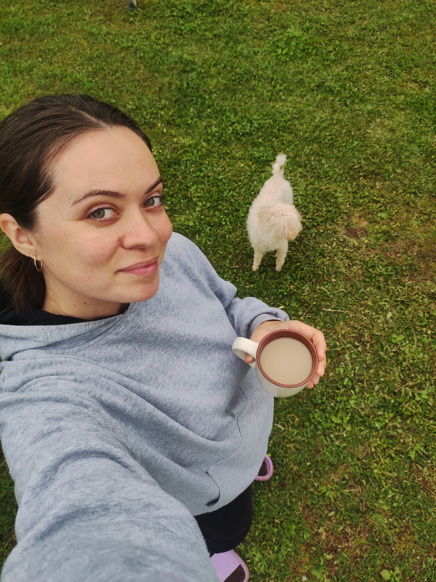 A high-angle selfie photograph depicts an adult female outdoors in a grassy area in the city of Foki, Russia. The woman has dark hair pulled back and wears a grey hooded sweatshirt over a black garment, black pants, and light purple slides. She holds a white mug with a brown rim, containing a light-colored beverage, in her right hand, extended towards the bottom right of the frame. She looks directly at the camera with a subtle smile. A small, cream-colored, curly-haired dog stands on the green grass behind and to the right of the woman, facing away from the camera and towards the upper right. The ground is covered in green grass with some visible patches of earth.