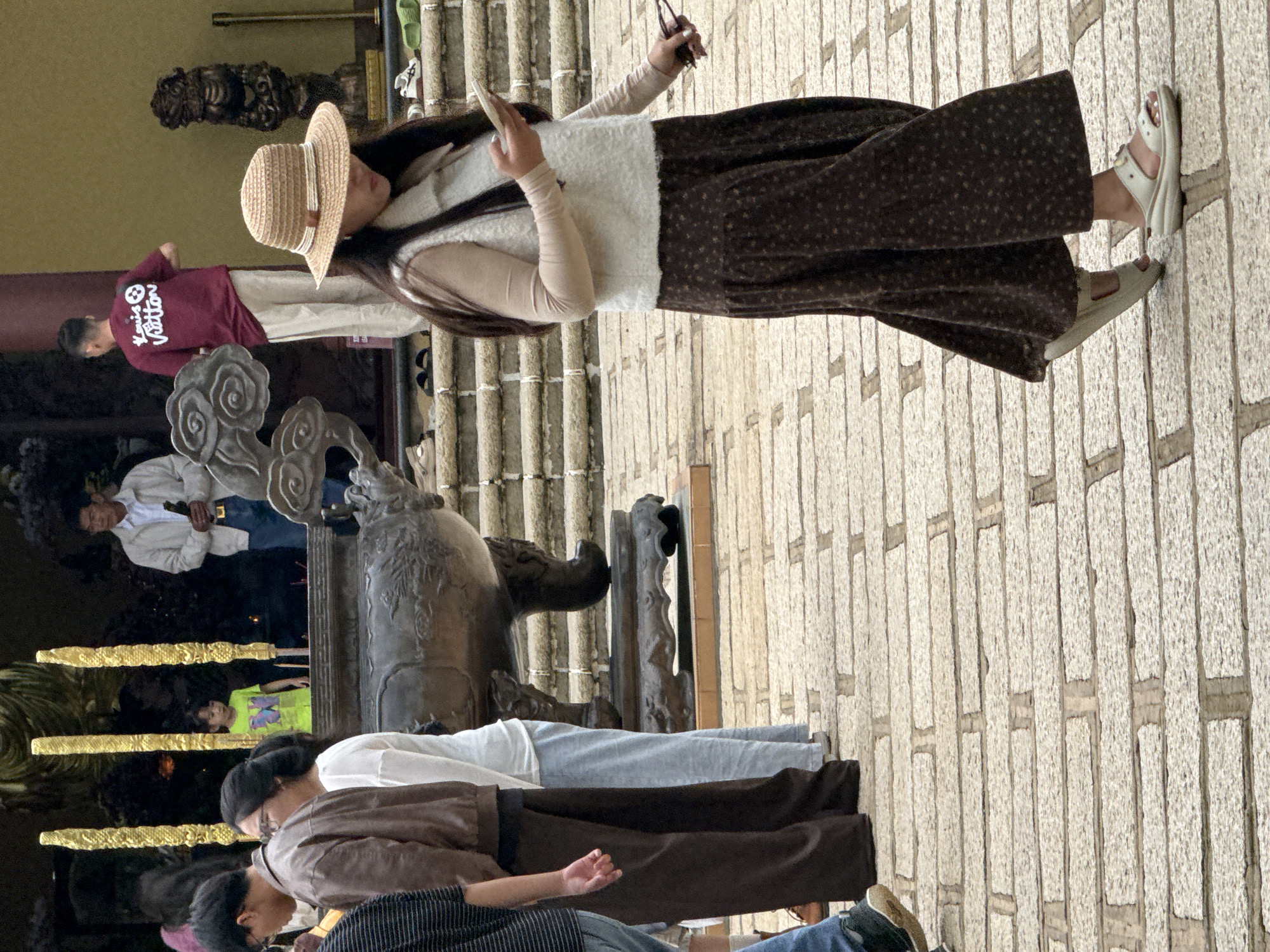 The image depicts a group of people standing on a cobblestone pathway in front of a building with a yellow facade and a large, ornate incense burner. The scene appears to be a temple or historical site, likely located in Da Lat, Vietnam, given the architecture and attire of the individuals.
In the foreground, a woman wearing a straw hat, a light-colored sleeveless vest over a long-sleeved beige shirt, and a dark floral skirt walks away from the camera, holding a smartphone in her right hand and sunglasses in her left. She is wearing white sandals.
To the left of the woman, a group of people are standing. One man wears a brown jacket and dark pants. Next to him is a woman in a white top and light blue pants. Further left, another man is visible, wearing a dark striped shirt and blue jeans.
In the background, an older man in a light jacket and jeans holds a dark object, possibly a bottle or a prayer item. Behind him, a young child in a bright green t-shirt stands near a large incense burner. Another individual, wearing a maroon t-shirt with white lettering and light-colored pants, is also in the background.
The environment includes stone steps leading up to the building, large decorative golden poles, and a carved statue to the right of the entrance. The pathway is made of light-colored cobblestones. The lighting suggests it is daytime, with some shadows indicating natural light.