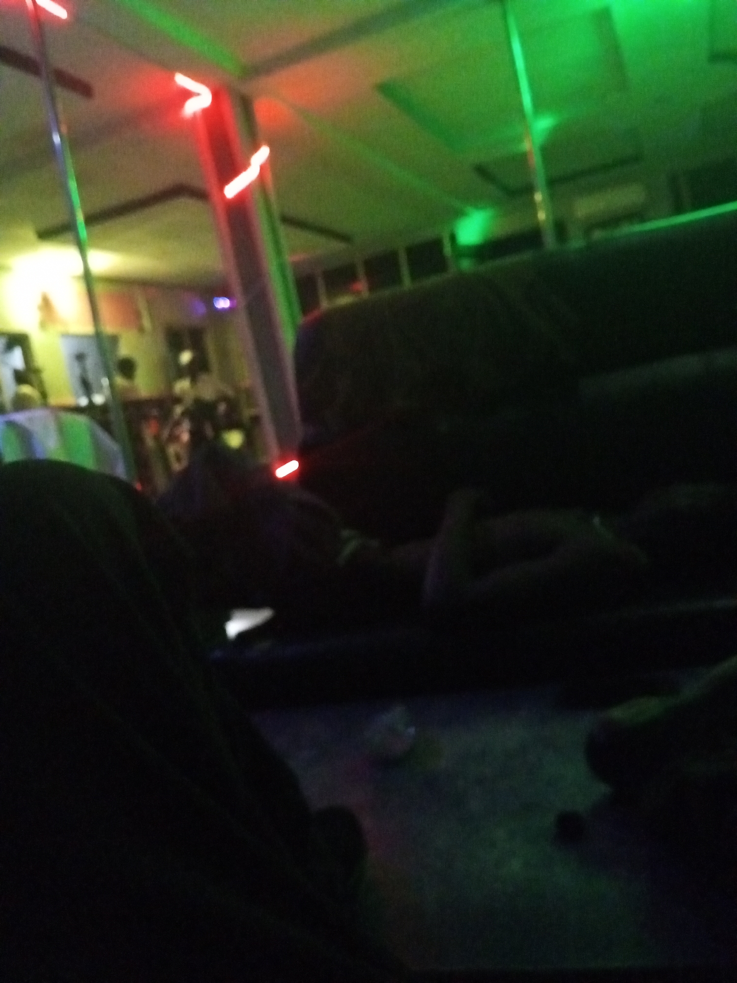 The image captures a dark, blurry indoor scene, characteristic of a bar, club, or lounge, illuminated primarily by green and red colored lighting. The ambiance suggests a nighttime setting.
In the immediate foreground on the left, the dark, indistinct form of a person's leg and lap is visible. A dark, reflective surface, possibly a table, occupies the bottom center, showing faint green reflections.
The middle and background feature dark, upholstered seating, likely couches or booths, where several blurry, indistinct figures are seated or reclining. To the left, a slender metallic pole extends from floor to ceiling, with colorful reflections along its length. Another, thicker column or pole is visible further back.
The ceiling and upper walls are lit with prominent green light strips, casting a distinct green hue over the upper half of the image. Bright red glowing elements, possibly LED strips or neon signs, are also scattered throughout, particularly on the right and near the columns. In the far background, areas are dimly lit with warmer yellow or orange tones, outlining what might be bar counters or shelves. A small blue light is also visible in the distant background.
The figures are too indistinct to determine specific activities, but they appear to be gathered in a social setting. There is no visible text in the image. The overall impression is one of a lively, albeit dimly lit, entertainment venue.