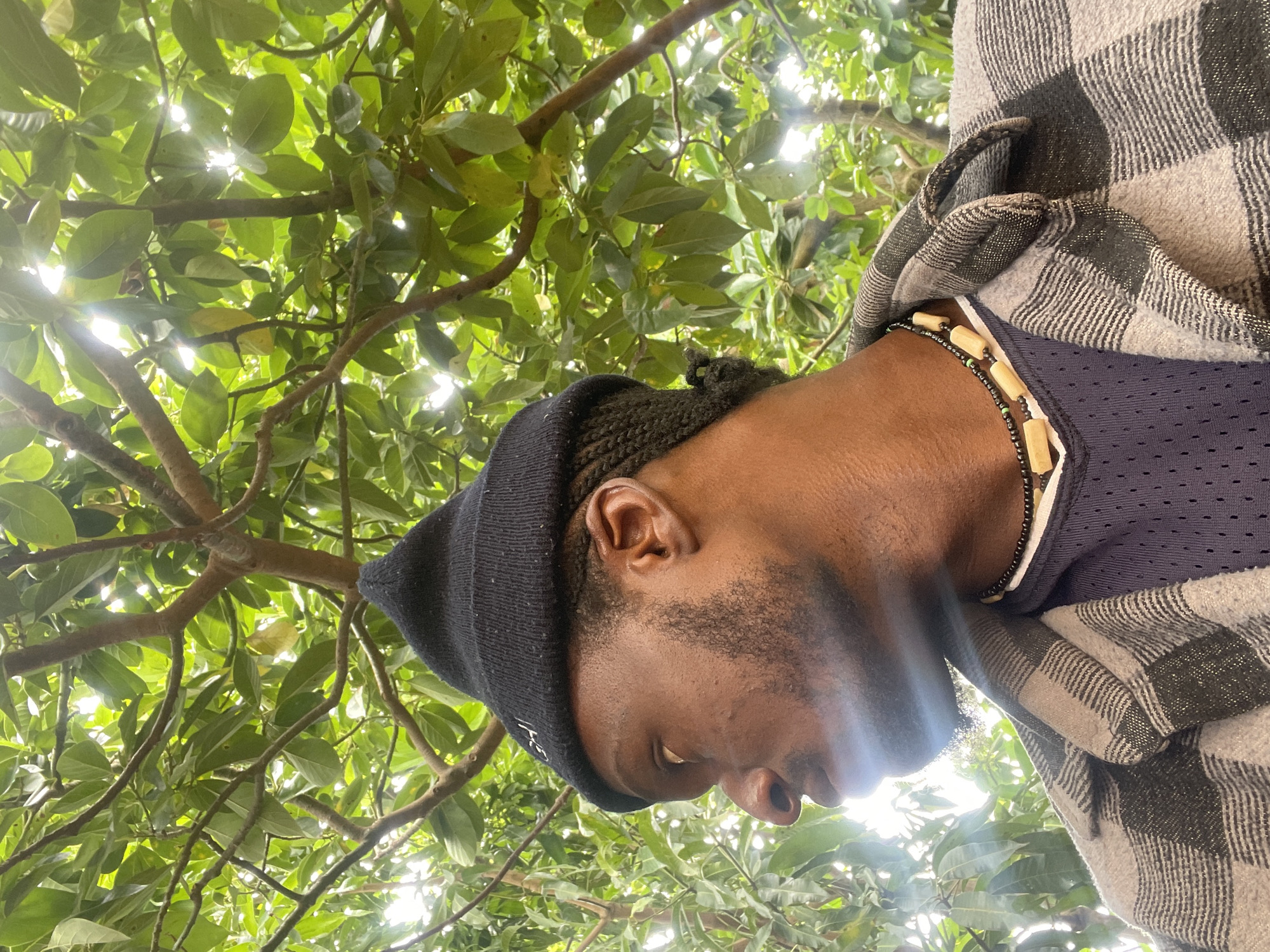 The image, rotated 90 degrees clockwise, captures a close-up, low-angle perspective of a person's profile against a lush green tree canopy. The subject, likely male with dark skin, is seen from the mid-chest upwards, looking downwards. He wears a black knit beanie that covers his head, revealing his ear and dark braided hair on the side. His jawline and chin show stubble or a short beard. He is dressed in a dark blue mesh top, over which he wears a grey and black plaid shirt or jacket, appearing to be of a soft or fleece-like material. A necklace with small black beads and larger, oblong, light brown wooden beads is visible around his neck.
The background is dominated by dense, vibrant green leaves and branches of a tree, with bright sunlight filtering through the foliage, creating natural highlights and lens flares. This indicates a clear, sunny daytime setting. The camera angle is from below, looking up at the person and the canopy. No specific activity is depicted; the person appears to be in a reflective or relaxed pose. The natural outdoor environment with bright sunlight is consistent with the specified location context of Jos, Nigeria. Faint, unclear white text is visible on the front of the black beanie, but its content cannot be deciphered.
