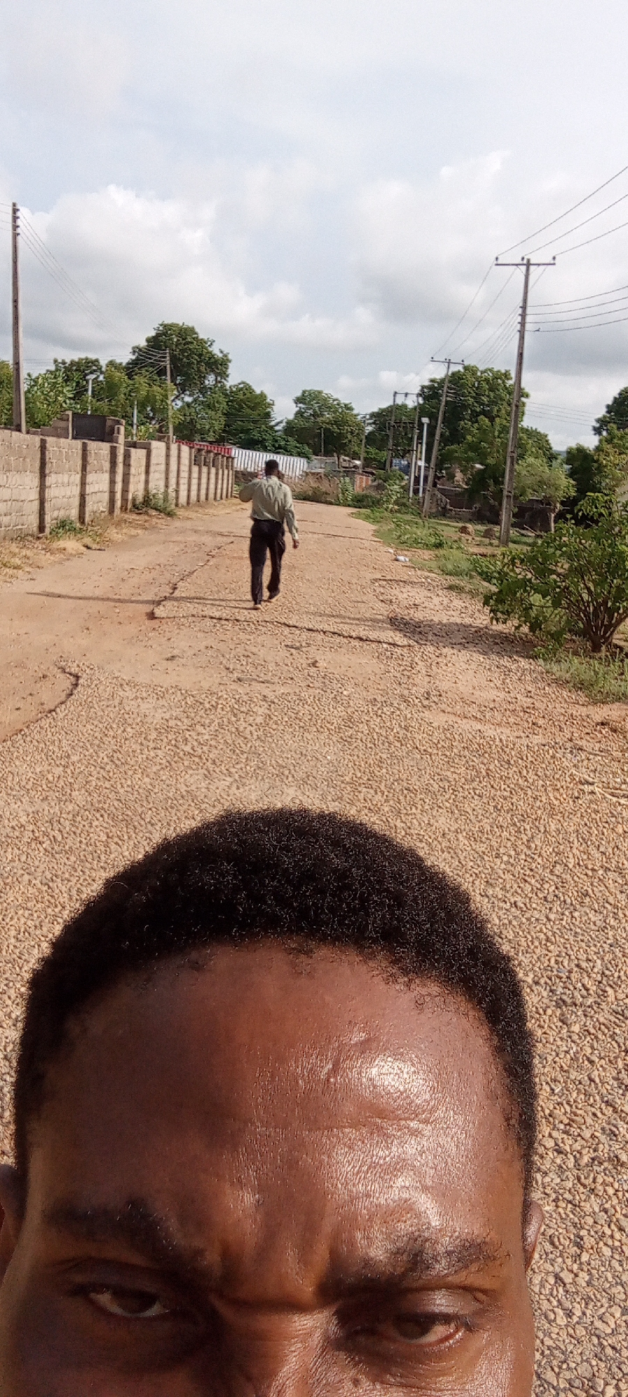 An eye-level perspective photograph depicts a gravel road in Adankolo, Nigeria. In the foreground, the upper portion of a man's face, including his forehead and eyes, is visible. Further down the road, a man wearing a light-colored shirt and dark trousers is walking away from the camera. A low concrete wall runs along the left side of the road, and utility poles with overhead wires are visible along the right side. Trees and other vegetation are present in the background. The sky is overcast with clouds.