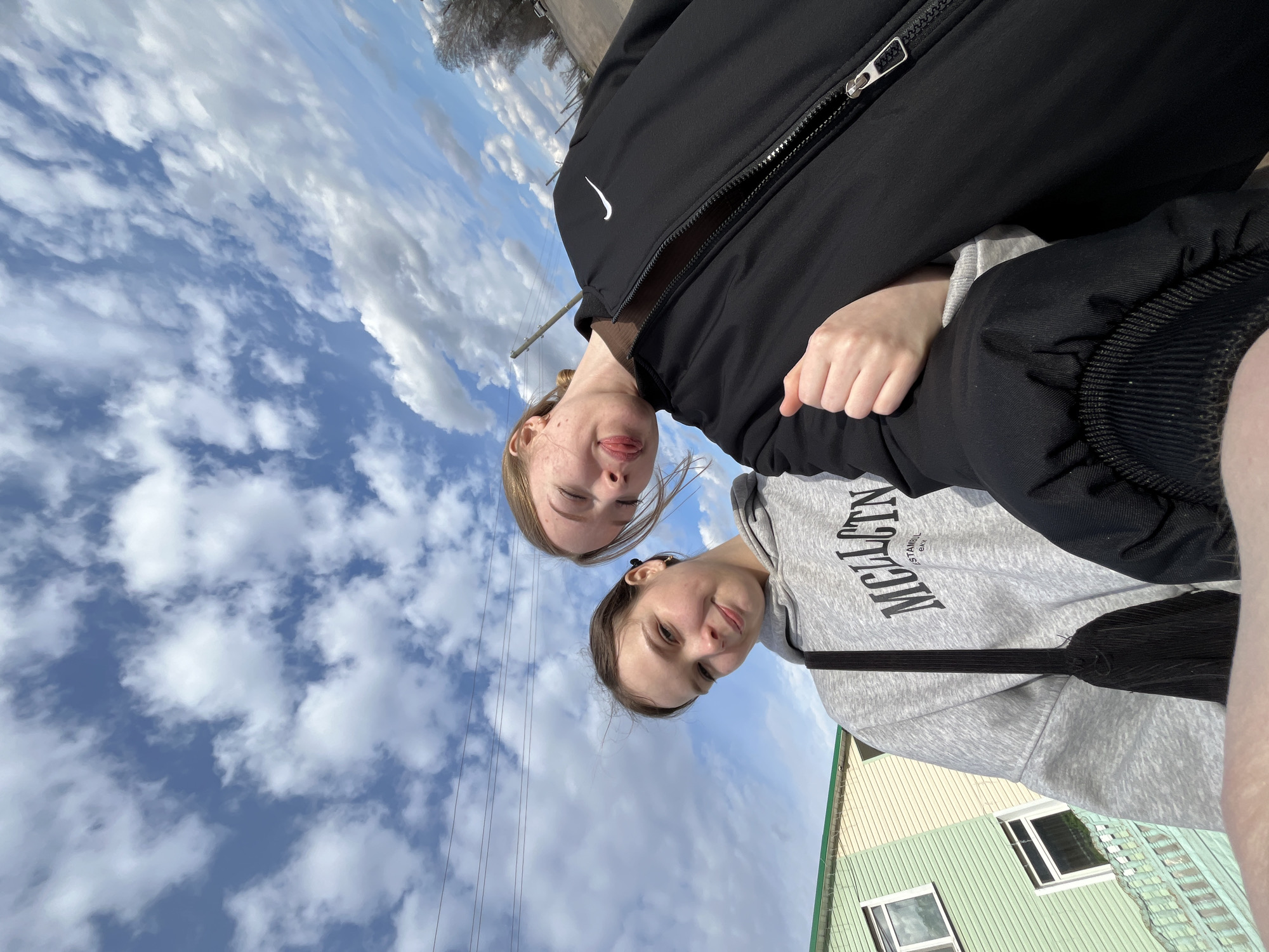 Two young women are captured in a low-angle outdoor shot under a bright, partly cloudy blue sky. The image is rotated, showing them looking down at the camera.
The woman positioned higher in the frame wears a black zip-up jacket, prominently displaying a white Nike swoosh logo on her left chest. She has light-colored hair, and her eyes are closed with her tongue playfully extended.
The second woman, slightly lower in the frame, wears a grey hooded sweatshirt with black text. The visible text includes "MCLCTN" with "ISTANBUL" and "EAN" printed beneath it. She has darker hair pulled back and offers a subtle smile to the camera. Her right hand rests on the arm of the other woman.
The background primarily consists of a vibrant blue sky dotted with scattered white clouds. Thin black power lines are visible stretching across the sky. To the right, a portion of a building with light green and light yellow siding and visible windows adds context to the scene, suggesting a residential or suburban setting in Sosnovo, Russia. The bright lighting indicates it is daytime. The overall mood of the image is casual and friendly, capturing a shared moment between the two.