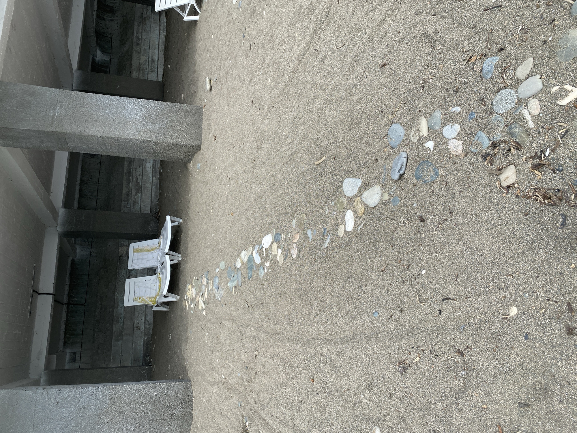 Two white plastic lounge chairs with yellow and white patterned cushions are set up on a sandy area underneath a concrete structure supported by large columns. A winding path of grey and white stones is laid out on the sand, leading from the foreground towards the lounge chairs. The environment appears to be a beach or a recreational area in Sokhumi, Abkhazia. The concrete structure suggests an outdoor, covered seating area. The sand shows some signs of being raked or disturbed, creating faint lines. There are scattered small pieces of driftwood and debris on the sand. No people are visible in the image.