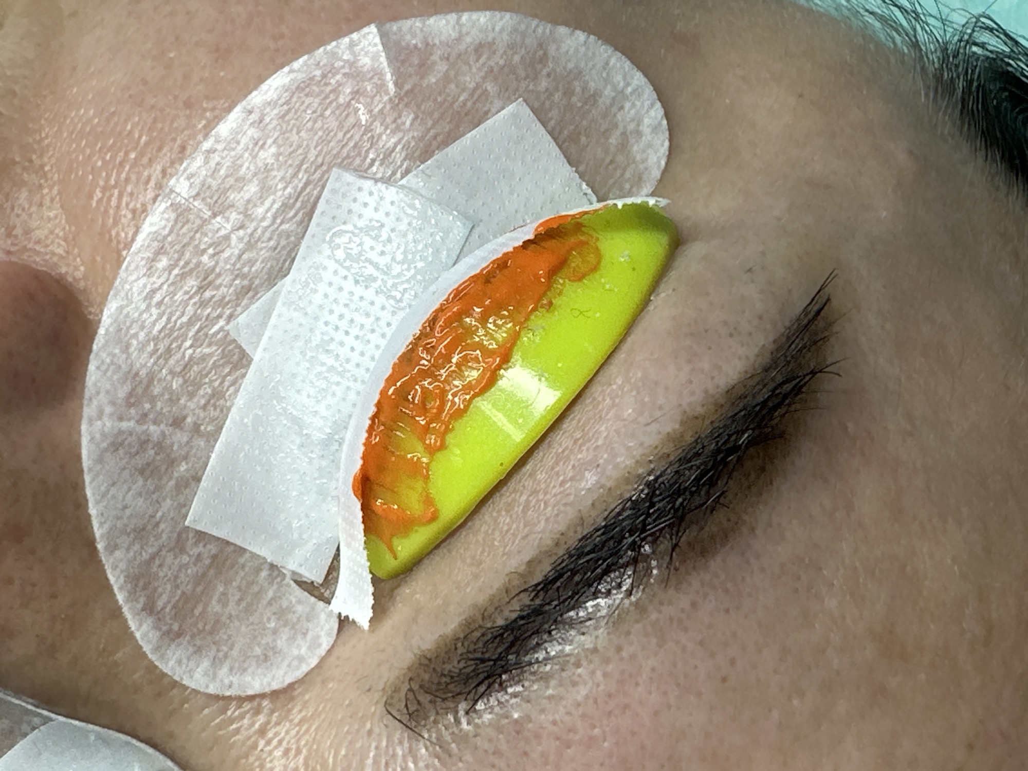 Close-up of a person's eyebrow area. An eyebrow is visible, with orange dye applied on a green tool resting adjacent to the brow. The area around the eye is covered with white absorbent pads and strips. Some upper eyelashes are also visible. The scene appears to be part of a cosmetic or beauty treatment. The context suggests it could be in Budva, Montenegro, based on the request, but that cannot be visually confirmed from the image itself.