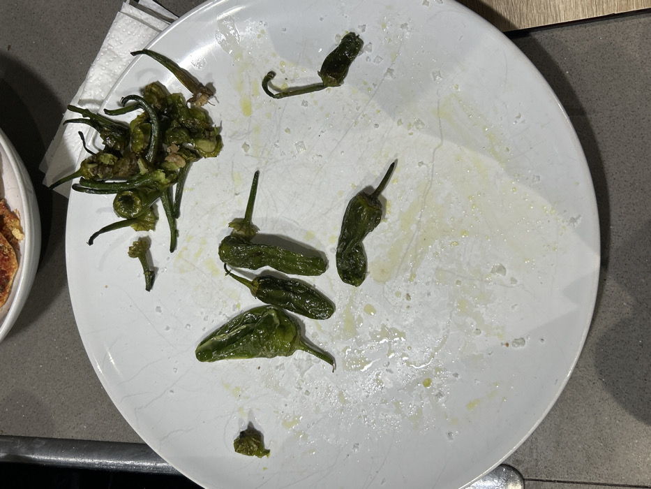 A white plate contains remnants of a meal, primarily comprising Padrón peppers. Some peppers are clustered together, seemingly fried, while others are scattered across the plate in varying states of consumption. The plate exhibits oil and salt residue. A napkin is visible to the left of the plate. A partial view of another dish is seen on the left side. The plate rests on a counter surface.