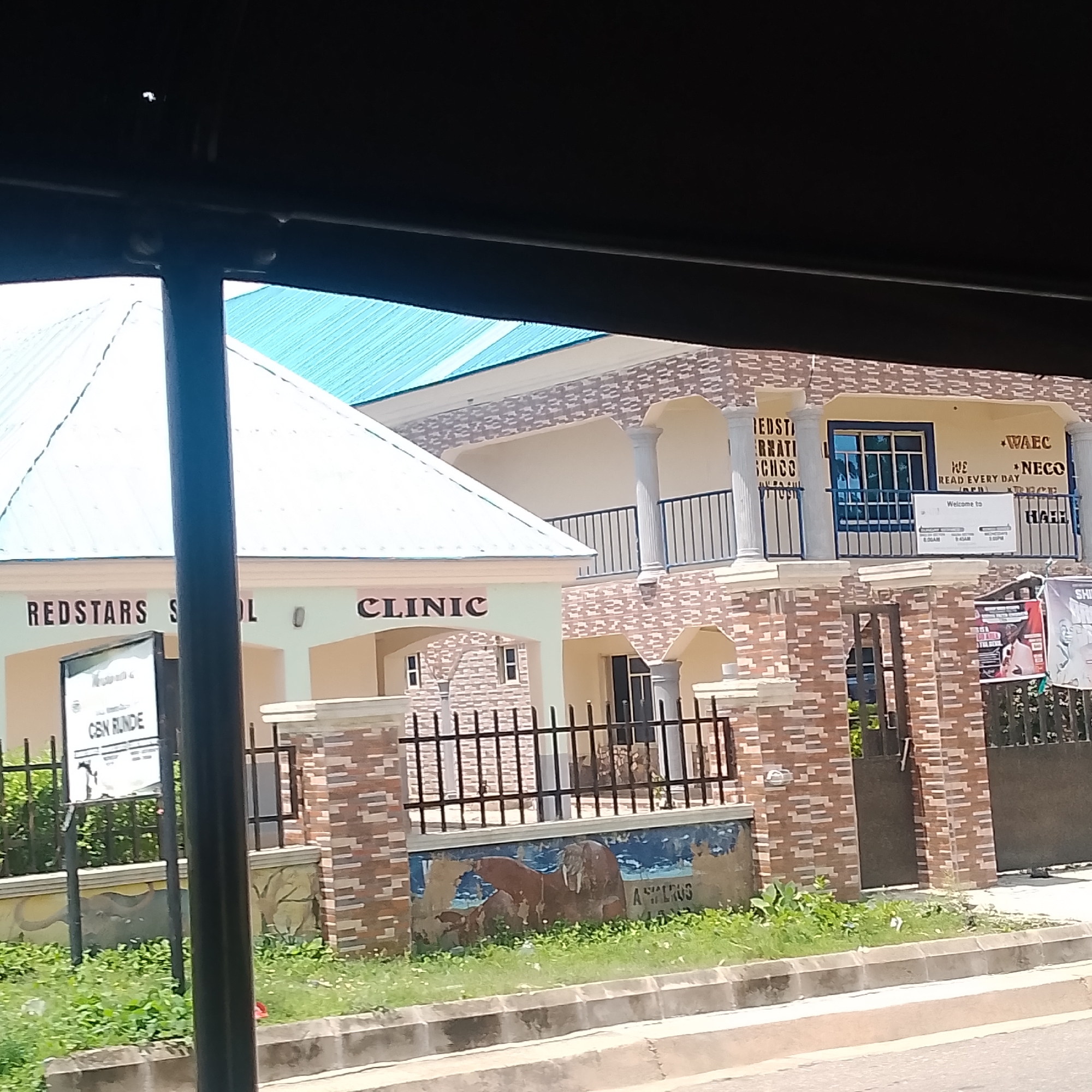 This media file shows a street view in Jalingo, Nigeria. The primary subjects are buildings, including a school and a clinic. The school, named "REDSTARS SCHOOL," features a blue-tiled roof and brick accents. Visible text on the school indicates it is a "REDSTARS S LO CLINIC" and also displays "WAEC" and "NECO," suggesting it's an educational institution preparing students for examinations. A sign near the entrance reads "CEN RUNDE." The clinic is adjacent to the school.
The scene appears to be an outdoor setting during daylight hours. The weather is not explicitly indicated but the bright lighting suggests it is likely daytime. There are no people prominently visible in the frame, with the focus being on the architecture and signage. The environment includes some greenery and a sidewalk, indicating a developed urban or semi-urban area. A mural depicting animals, possibly elephants, is painted on a wall. The overall impression is that of a functional community facility.