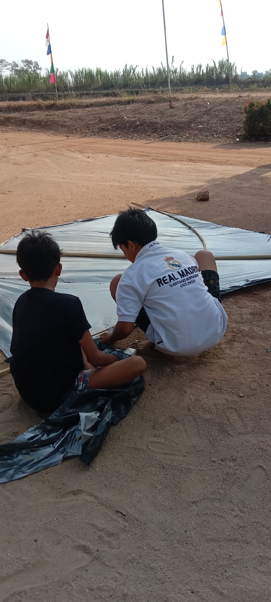 In the city of Utama Jaya Mataram, Indonesia, two boys are on the ground preparing a large, dark plastic sheet. One boy, wearing a black t-shirt and patterned shorts, is seated with his back to the camera. The other boy, wearing a white polo shirt with "REAL MADRID" printed on the back, is kneeling and appears to be working with the plastic sheet. The ground is sandy and dry, with some vegetation visible in the background, including what appears to be a field of tall grass or reeds. Two flagpoles with colorful flags are visible in the distance, suggesting a festive or event setting. The overall environment is outdoors in a rural or semi-rural area.