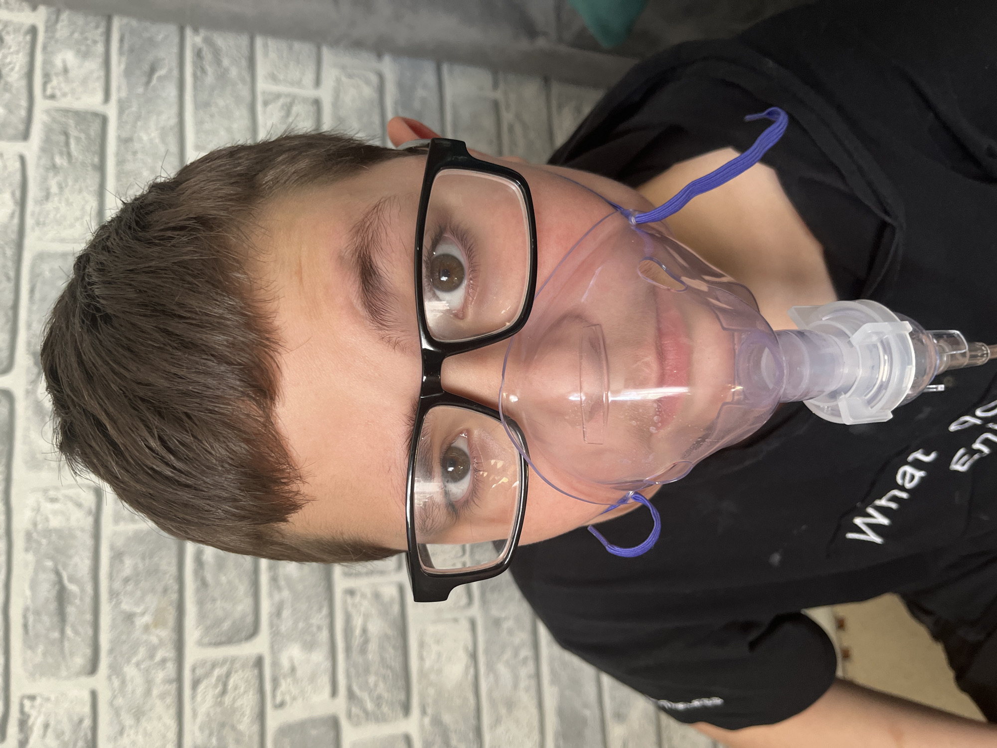 A young person, likely a child or adolescent, is depicted in a close-up, chest-up shot. They are wearing a clear plastic nebulizer mask over their mouth and nose, with blue elastic straps visible around the back of their head. Black-rimmed glasses are worn over their eyes, which are open and looking directly towards the camera. The person has short, dark brown hair and a light complexion.
They are wearing a black t-shirt, and partially visible white text on the shirt reads "What" and the start of another word, possibly "En". The background features a light-colored wall with a textured, brick-like pattern, suggesting an indoor setting. The image primarily focuses on the person's face and upper torso, with the medical device being a prominent feature. The activity shown is the use of a nebulizer, indicating a medical or therapeutic treatment. The person's expression is neutral. The provided location context for this image is Vesyoloye, Russia.