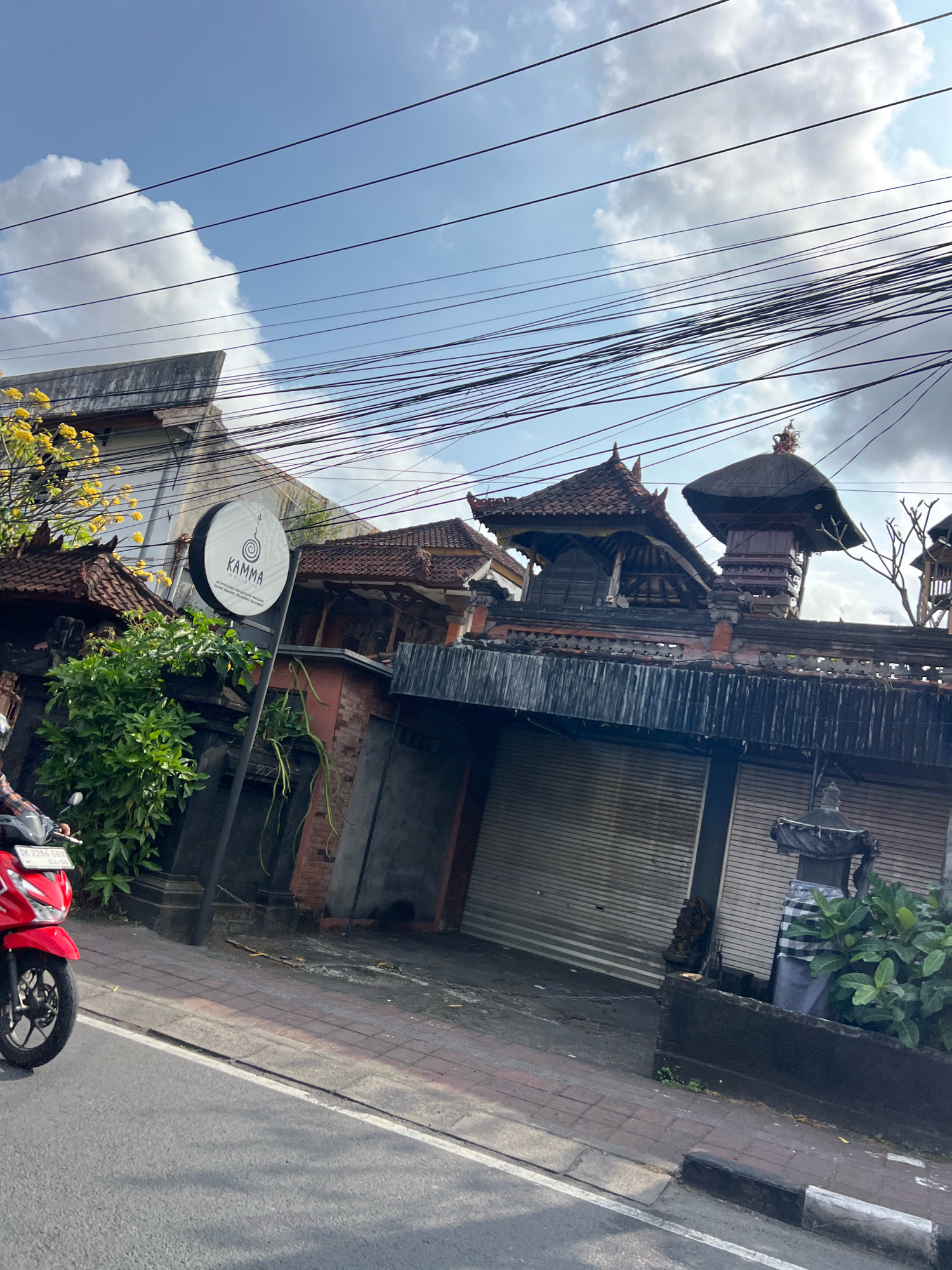 In Denpasar, Indonesia, a street view captures a building with traditional Balinese architecture, characterized by tiered, dark-tiled roofs and intricate wooden details. A prominent circular sign with the text "KAMMA HOUSE OF MASSAGE" is mounted on a pole in front of the structure. The building appears to be a spa or wellness center. A red motorcycle, partially visible on the left, is in motion on the paved road. The environment features lush green foliage, and an overhead network of power lines crisscrosses the sky, which is partly cloudy. A statue dressed in a checkered cloth stands near the entrance of the building. The building's facade is lined with rolled-down metal shutters, suggesting it may be closed or preparing for closure.