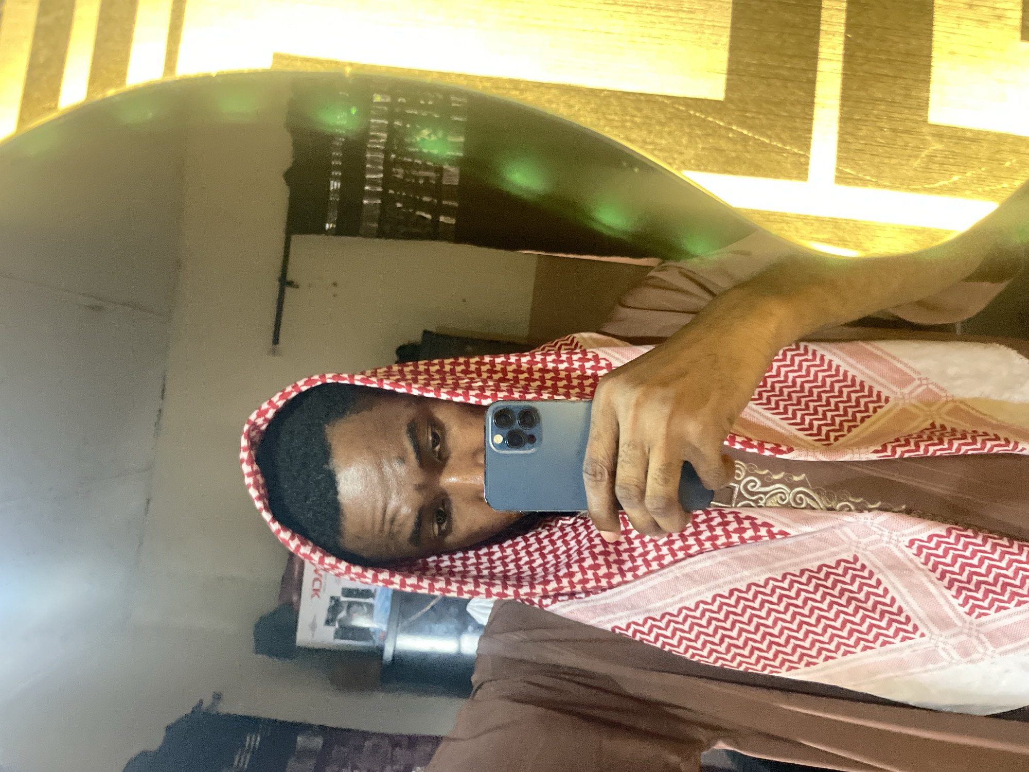 A man wearing a red and white patterned headscarf is taking a selfie in a mirror. He is holding a blue smartphone. The background is blurry and shows a light-colored wall with a patterned surface. The location cannot be determined from the image.