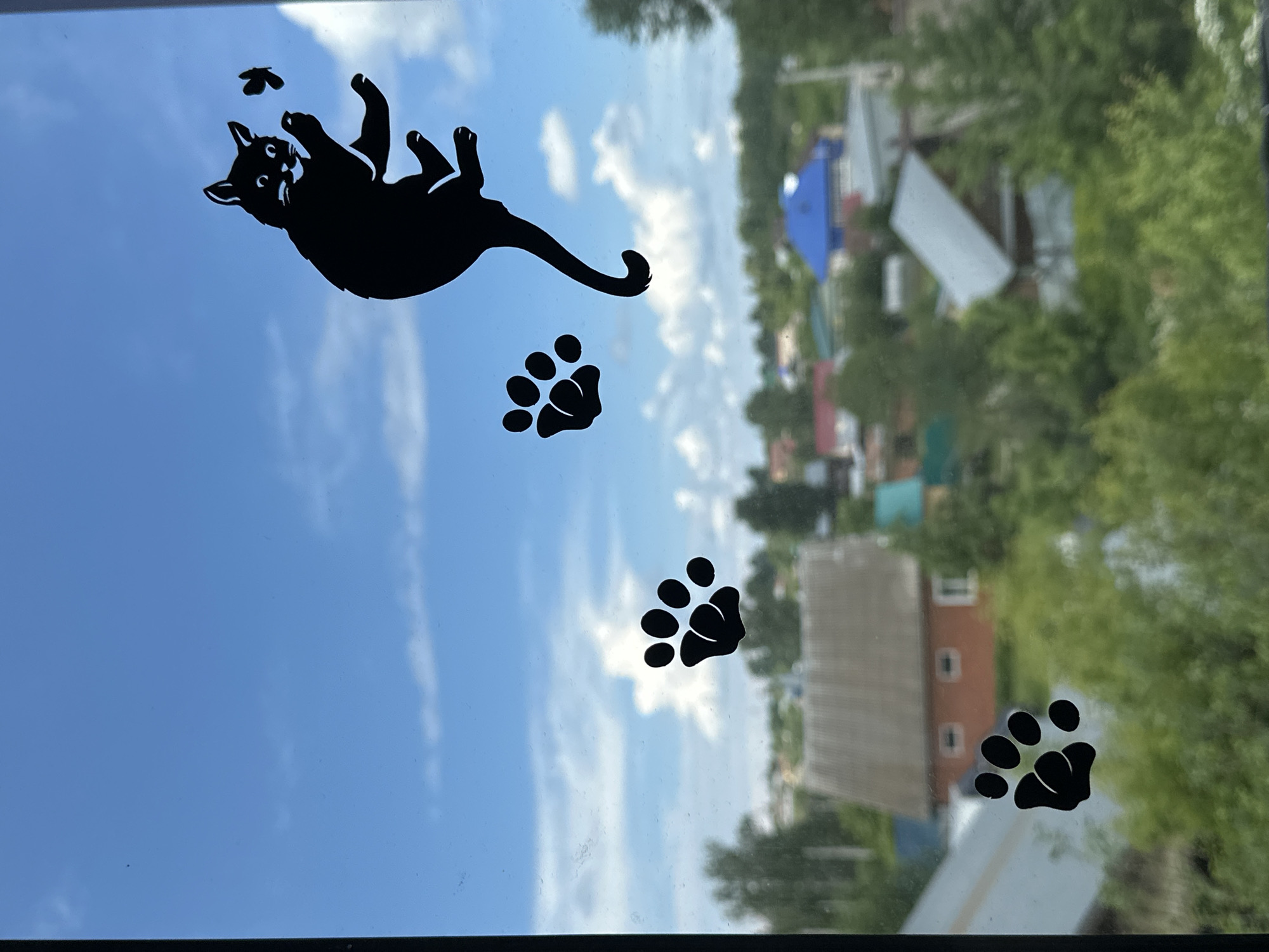The image shows a window with decorative black cat and paw print decals. The cat decal is in the upper left and is depicted as if it's falling or playing with a small fly. Three paw print decals are spaced below the cat. The background is a view through the window showing a residential area with houses, trees, and a blue sky with wispy clouds. The location is Unknown, Unknown.