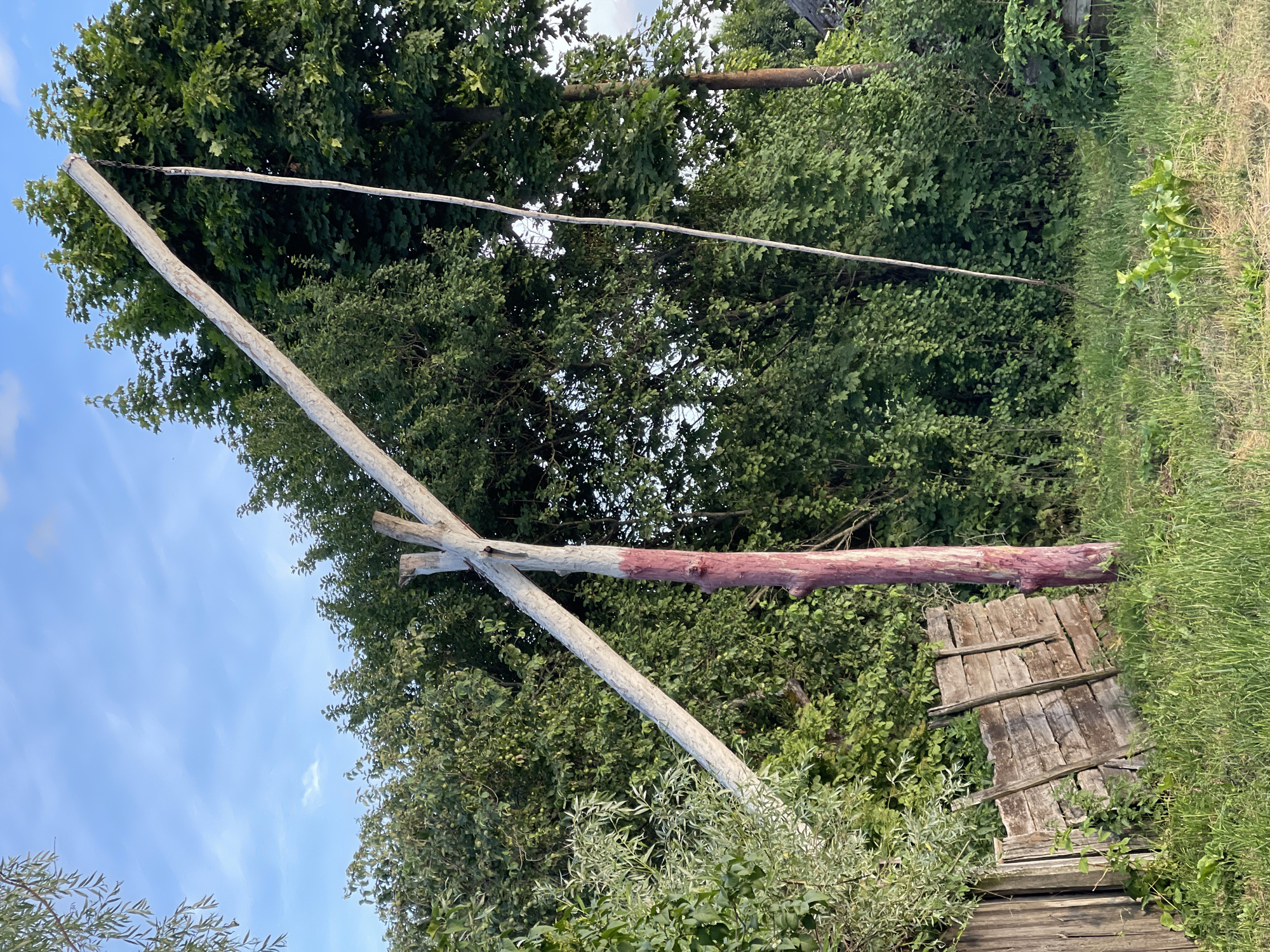 The image shows a rustic wooden structure in an outdoor, natural setting. The primary focus is a cross-shaped wooden beam supported by two upright poles. One of the horizontal beams has a reddish hue, possibly from paint or a natural stain. Below this structure, a weathered wooden platform, partially overgrown with vegetation, is visible.
The background is filled with lush green trees and foliage, suggesting a wooded area or a garden. A clear blue sky with scattered wispy clouds can be seen above the trees, indicating daytime, likely clear weather. The overall impression is one of an old, possibly abandoned, piece of rural infrastructure. There are no people visible in the image, and no text is present. The scene evokes a sense of tranquility and the passage of time.