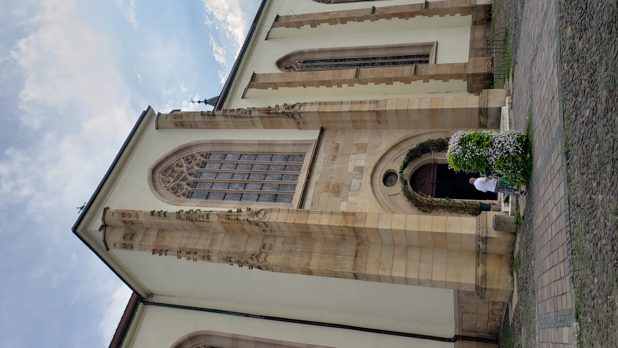 An external, eye-level, wide shot captures the facade of the St. Nicholas Cathedral in Prešov, Slovakia. The creamy yellow stone building features gothic architectural elements, including pointed arched windows with intricate tracery. A man, wearing a white t-shirt and plaid shorts, is standing in front of the cathedral entrance, which is adorned with a floral archway and a large, cone-shaped planter overflowing with white and pink flowers. The ground in front of the cathedral is paved with cobblestones and decorative brickwork. The sky above is partly cloudy, with a few scattered white clouds against a light blue background. The overall scene suggests a serene and well-maintained exterior of a historic church.