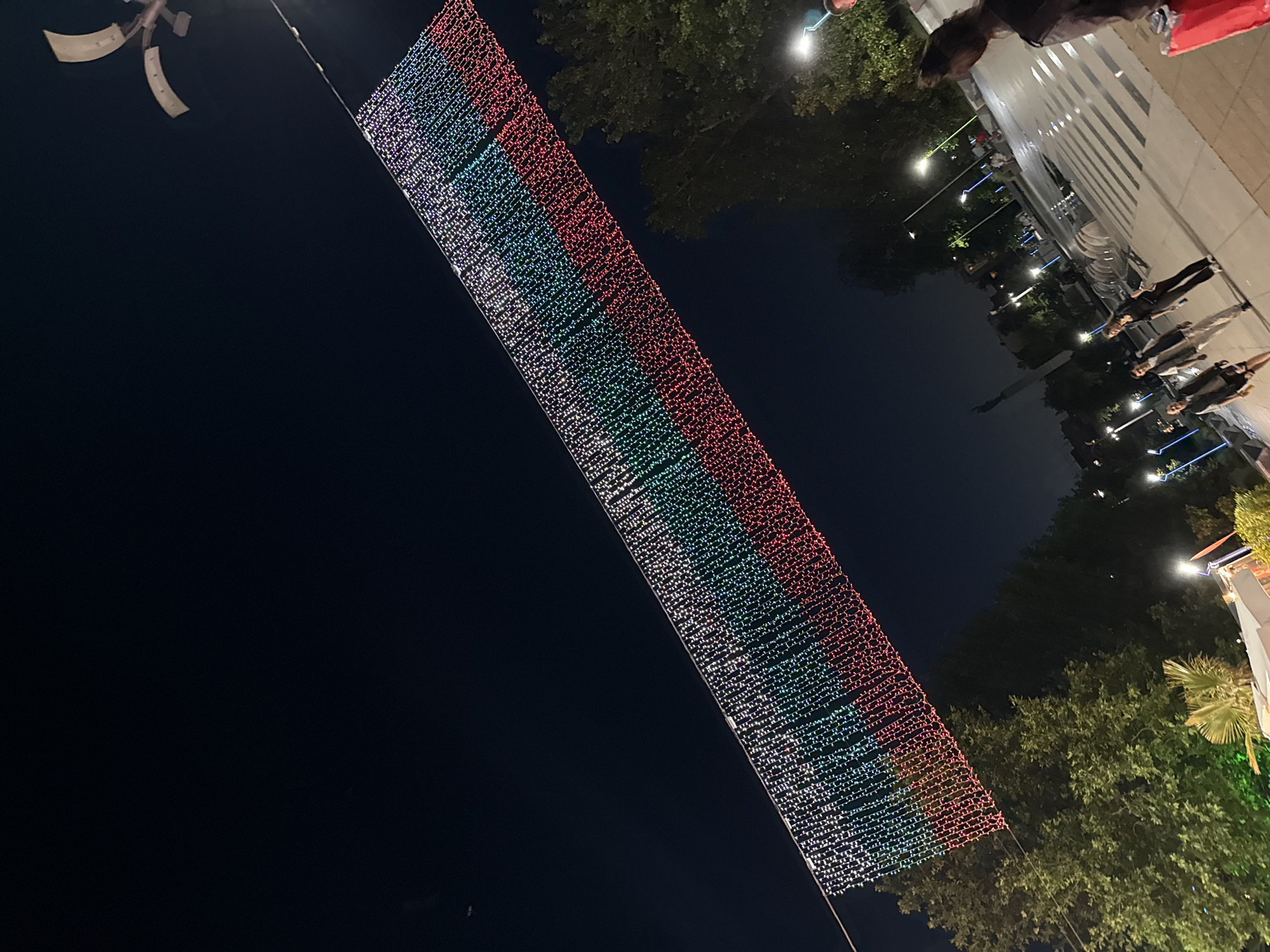 At night, a large string of lights resembling the Bulgarian flag hangs diagonally across the frame, suspended between two unseen points. The lights are arranged in three horizontal bands: white at the top, green in the middle, and red at the bottom, mimicking the national flag. Below this festive display, an open plaza is visible. Several people are walking across the plaza, some appearing to be in conversation. Streetlights with blue accents illuminate the area, and trees line the perimeter of the plaza. In the background, buildings and a distant monument can be seen. The scene is set in the city of Burgas, Bulgaria.