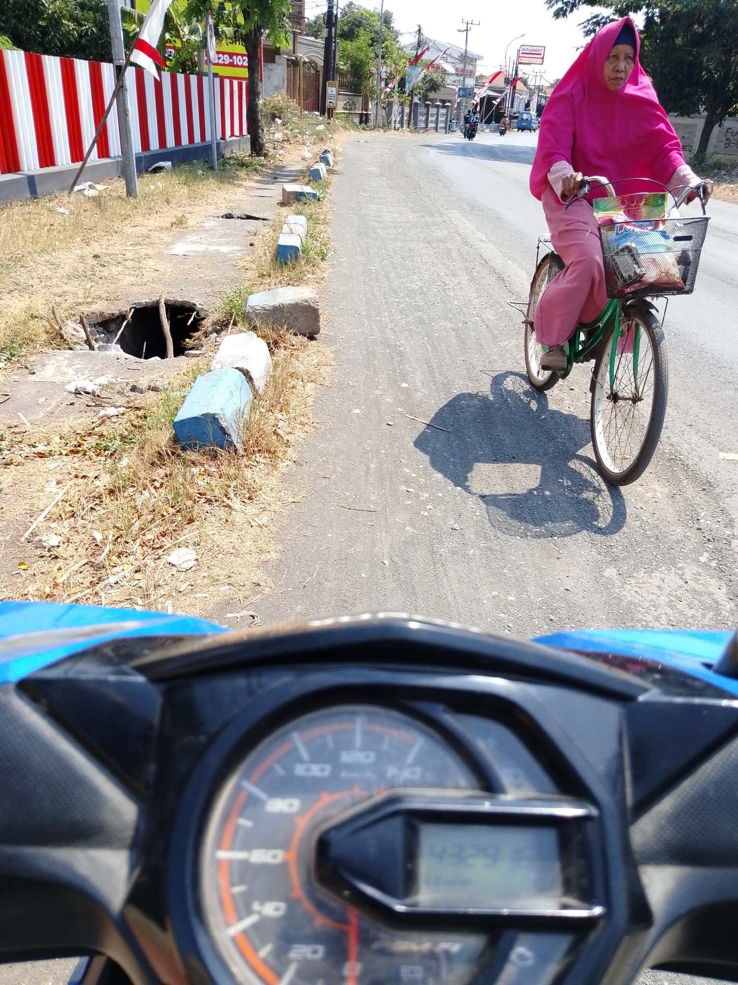 In Pasuruan, Indonesia, a woman in a pink hijab and matching pants rides a green bicycle on a paved road. The bicycle has a basket on the front containing various items. The road is lined with a dry, grassy verge and a sidewalk. On the left side of the road, there is a red and white striped fence and a large hole in the ground next to the sidewalk. The sky is bright and sunny, casting a shadow of the woman and her bicycle on the road. The foreground shows a partial view of a blue motorcycle's dashboard, indicating the time is 4:32.