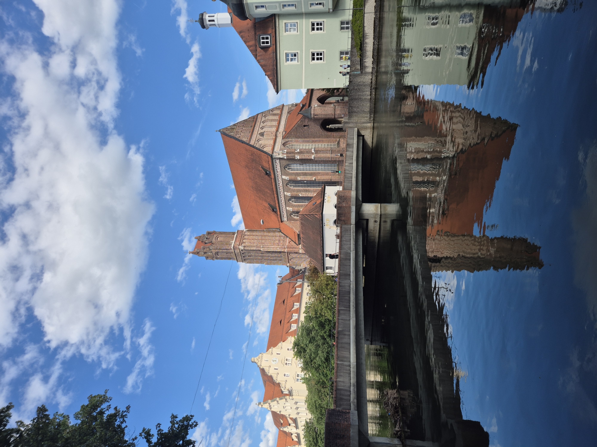 The image depicts a serene canal scene in Landshut, Germany, dominated by a majestic Gothic brick church with a tall tower, its orange-tiled roof gleaming under a partly cloudy blue sky. To the left, a pale yellow building with a similar red roof and a clock face is visible. On the right, a pastel green building with white-framed windows and a dark turret stands adjacent to the church. A concrete bridge spans the canal in the foreground, with the reflections of the buildings, bridge, and sky creating a mirrored image on the calm water. A few people are discernible on the canal bank and crossing the bridge, appearing as small figures engaged in casual strolling. The overall environment suggests a peaceful urban setting with a strong sense of historical architecture.