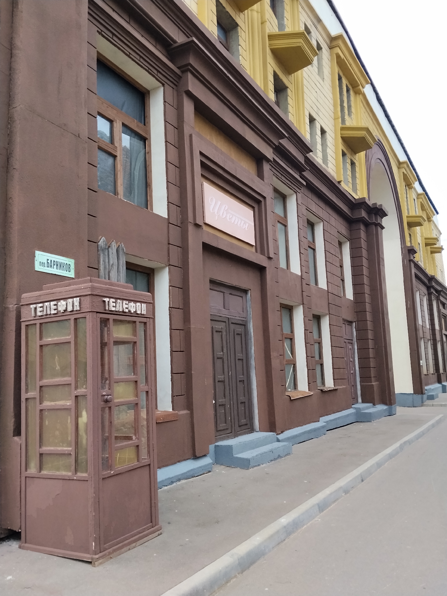 A view of a brown building facade with yellow accents and a street in front, possibly in Moscow, Russia. In the foreground, a weathered, brown telephone booth stands on the sidewalk. The booth has glass panels and is labeled "ТЕЛЕФОН" (TELEPHONE) in raised letters on multiple sides. A blue street sign that reads "пер. БАРНИКОВ" (Barnikov Lane) is mounted on the building above the telephone booth. The building itself features arched windows and decorative elements. A sidewalk curves away from the building and into the distance. The sky is overcast.