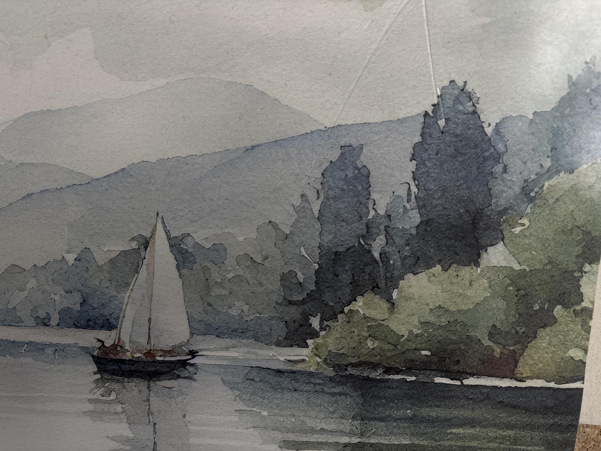 The image is a watercolor painting depicting a serene landscape scene.
Environment: The painting features a body of water in the foreground, likely a lake or river, with calm reflections. The middle ground consists of a forested area with varying shades of green and blue-gray, suggesting different tree types and depth. In the background, there are hazy mountain ranges, painted in lighter shades of blue-gray, creating a sense of distance.
Objects: A sailboat with white sails is prominently featured on the water. It appears to be moving gently across the calm surface. The painting is also made up of paper or wood, and there are some cork boards at the very bottom of the image.
The location is not confirmed to be Troyan, Bulgaria, as no specifics are visible in the photo.