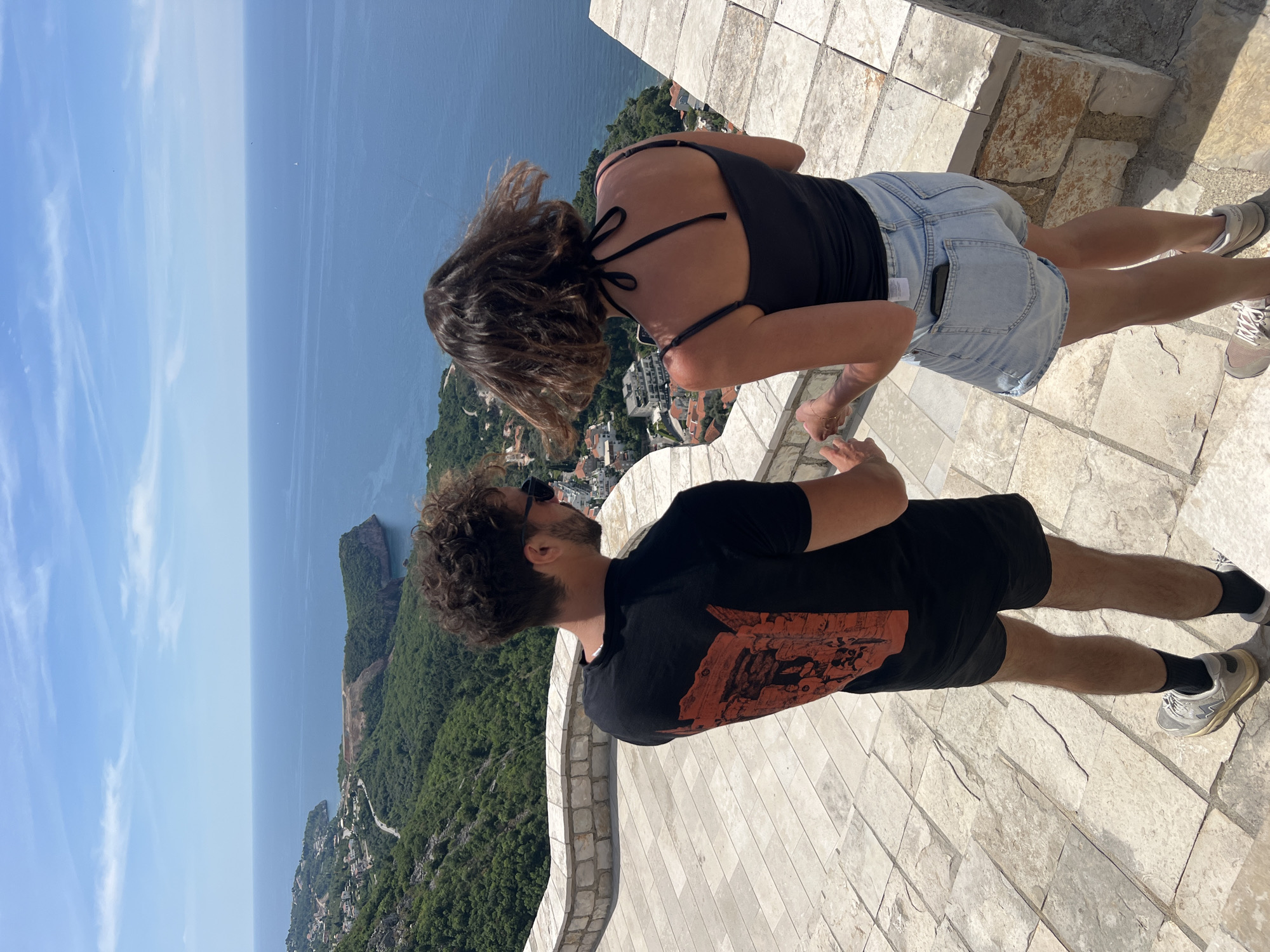 Two individuals, a female and a male, are standing on a stone terrace, possibly a high point with a view overlooking a coastline in Sveti Stefan, Montenegro. The female is facing away from the camera, with her hair blowing in the wind. She is wearing a black tank top and light denim shorts. The male is standing next to her, his back turned slightly towards the camera. He is wearing a black t-shirt with a large graphic design on the back and black shorts. The view from the terrace includes the sea, rocky coastal features, and green forested areas. In the distance, buildings and infrastructure characteristic of a coastal town can be seen. The sky is blue with scattered clouds. The terrace is paved with light-colored rectangular stone tiles.