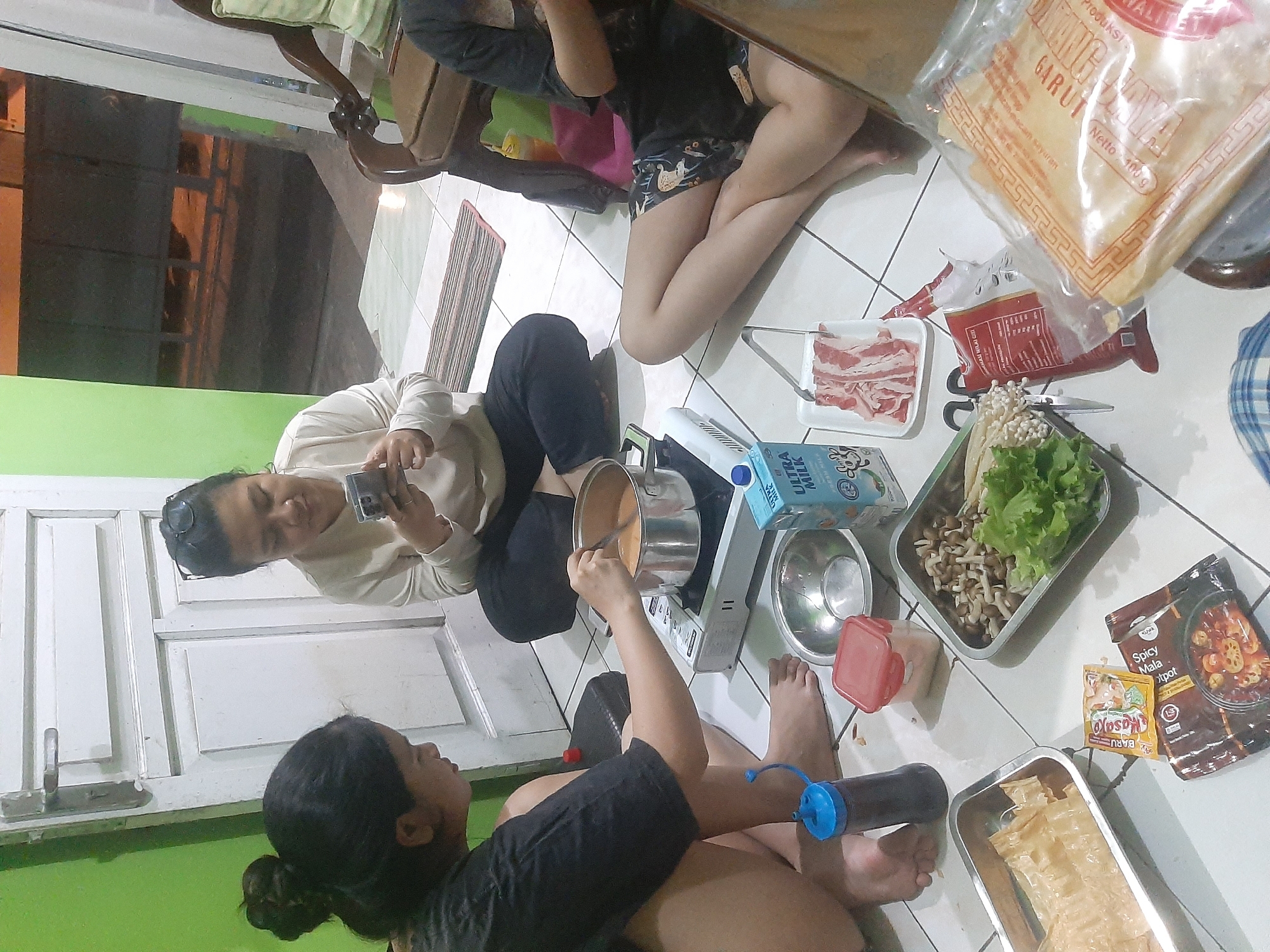 In Sumampir, Indonesia, a group of four individuals are gathered on the floor around a portable induction stove and various ingredients for a hot pot meal. The setting appears to be an indoor residential area, possibly a living room or dining area, with tiled flooring and a partially open door in the background. The walls are painted in light green.
Two women are prominently featured in the foreground. One woman on the left, with her hair in a bun, is stirring a pot of soup on the induction stove with a spoon. She is wearing a black t-shirt. Beside her, on the floor, is a dark bottle with a blue dispenser lid. Another woman in the center, wearing a light-colored long-sleeved shirt and glasses perched on her head, is looking at her smartphone. She is seated next to a carton of "ULTRA MILK."
To the right of the central woman, another person's legs are visible, wearing dark patterned shorts. This individual is reaching towards a plate of thinly sliced raw meat, presumably for the hot pot. Further to the right, a wooden chair with a light green cushion is partially visible.
On the floor, several food items are arranged. These include a metal tray filled with fried tofu sheets, a metal tray containing mushrooms and lettuce, a small red container, a small metal bowl with a white substance (possibly salt or seasoning), and packets of seasonings labeled "Kasjo" and "Spicy Mala Hot Pot." A large transparent package with red and gold Asian lettering is also present in the foreground. The overall scene suggests a casual and communal meal preparation.