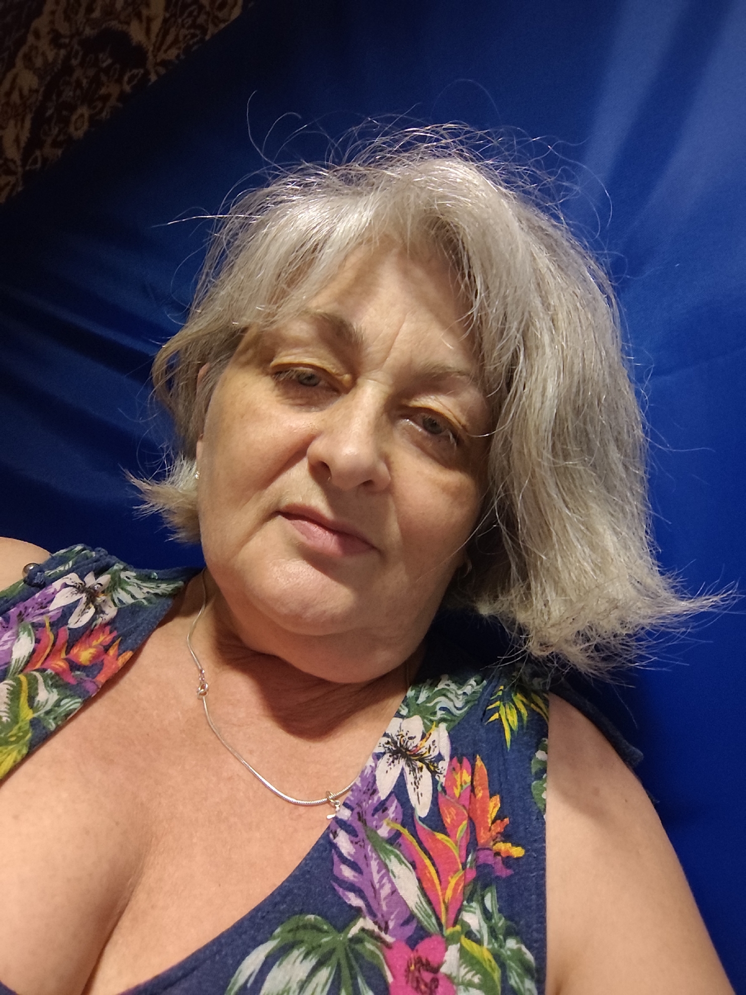 A middle-aged Caucasian woman with shoulder-length gray hair and blue eyes is pictured from the chest up. She is wearing a floral print top with a V-neck and a thin silver necklace. Her skin has wrinkles and sunspots. The background is a deep blue fabric, possibly a curtain or backdrop, with a patterned tapestry visible in the upper left corner. The lighting is soft and appears to be coming from the front. The image is a selfie, taken at a slight downward angle. The scene appears to be indoors, likely in a residential setting. The specific location in Krasnodar, Russia, cannot be determined from the image alone.