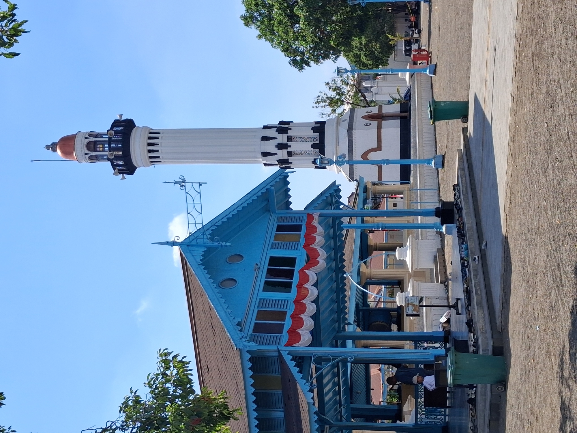 The image depicts the Menara Masjid Agung Surakarta, a historic mosque located in Surakarta, Indonesia. The tall, white minaret with black accents and a copper dome dominates the background. In the foreground, a brightly painted blue pavilion with a brown roof is visible, adorned with a red and white banner. Several pairs of shoes are neatly arranged on a raised platform in front of the pavilion, suggesting people are inside or have recently been. A green trash can with wheels stands to the left of the pavilion. Two individuals, one adult and one child, are partially visible within the blue structure. The ground is a mix of paved areas and gravel. The sky is clear and blue, with a few scattered clouds. Trees flank the scene on the right and left sides.