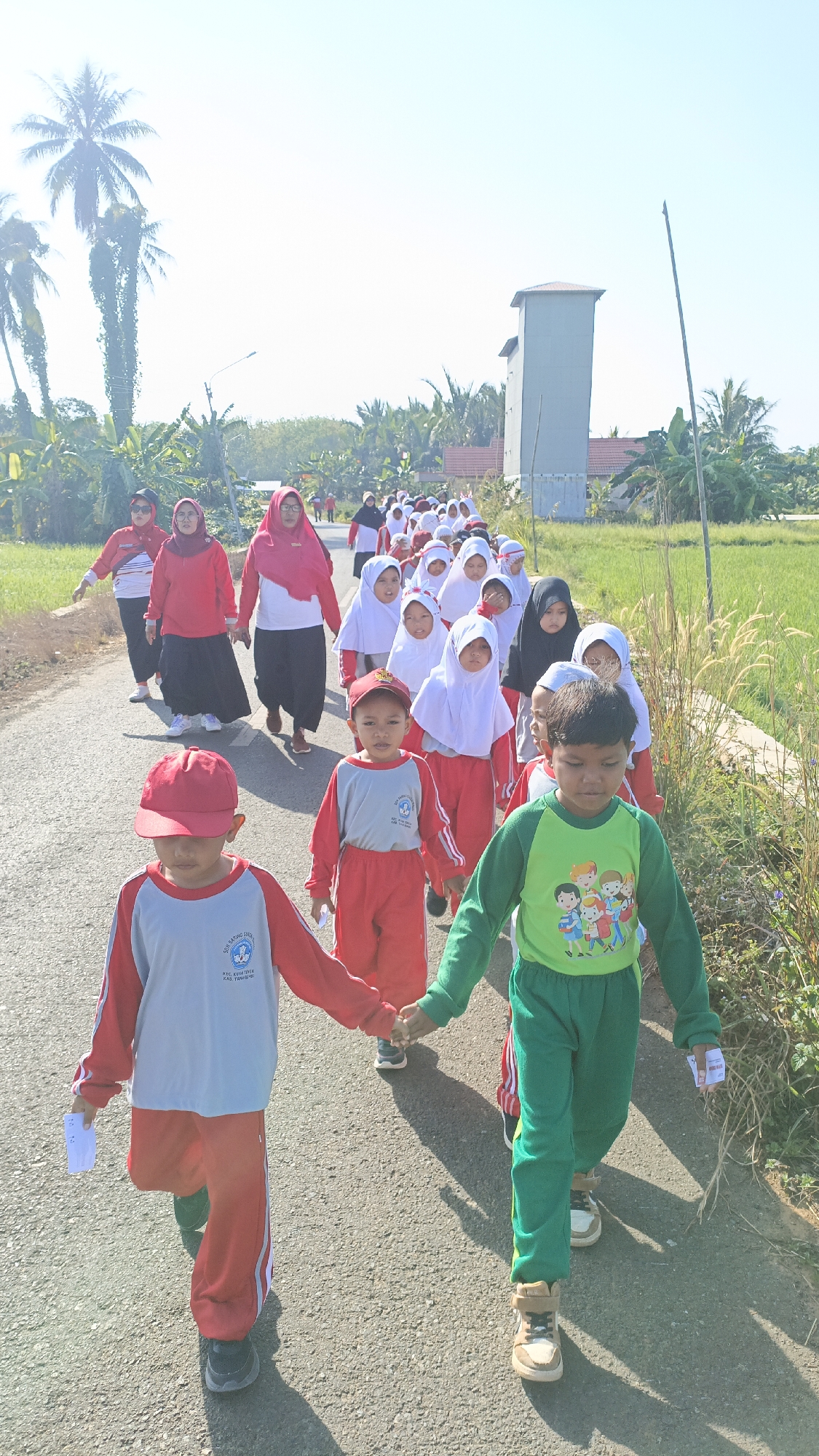 In Batarang, Indonesia, a group of young students in red and white uniforms, some wearing hijabs, walk along a paved road lined with vegetation and palm trees. The students are holding hands and appear to be participating in an organized activity, possibly a parade or a school outing. Two young boys in the foreground are wearing red caps and holding hands. One boy in a light grey and red uniform carries a piece of white paper. The other boy wears a green shirt with a colorful cartoon graphic and green pants. In the background, adults in similar red attire and hijabs are also walking. A grey tower-like structure is visible in the distance, along with rice paddies and rural houses. The sky is clear and bright, indicating a sunny day.