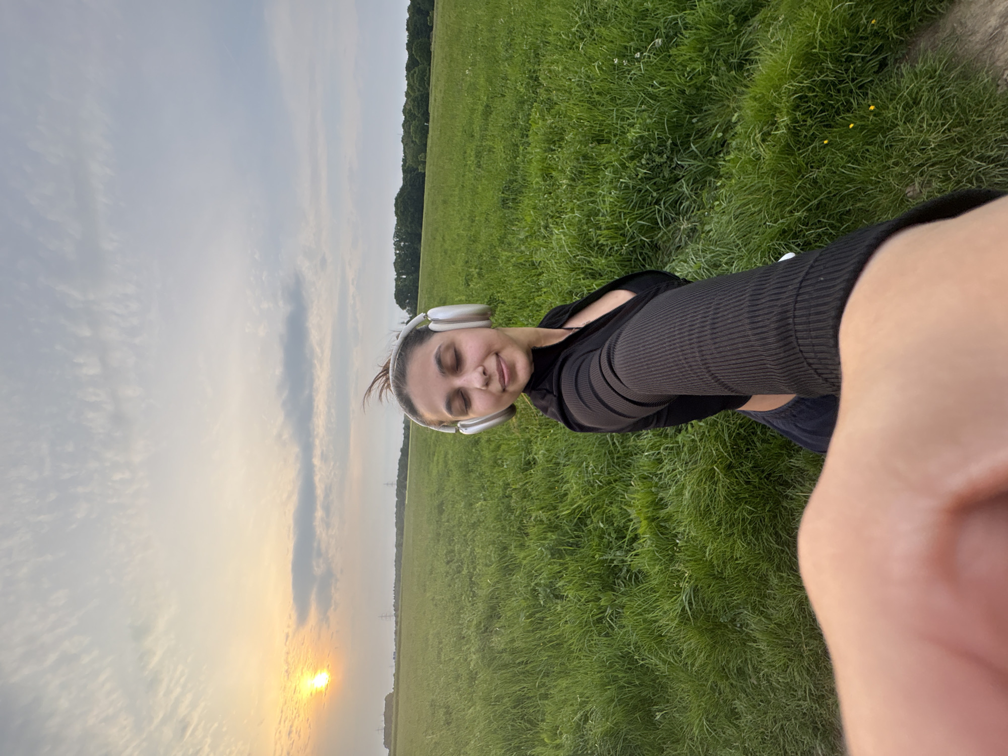 A young woman with headphones takes a selfie in a grassy field at sunset in Mönchengladbach, Germany. The scene includes a horizon line of trees in the distance, and a sky with cloudy patterns and a prominent sun setting on the left side of the frame. The woman is wearing a dark top with mesh sleeves. The field has dense green grass with patches of lighter green areas. Part of the selfie taker's arm is visible in the lower right corner.